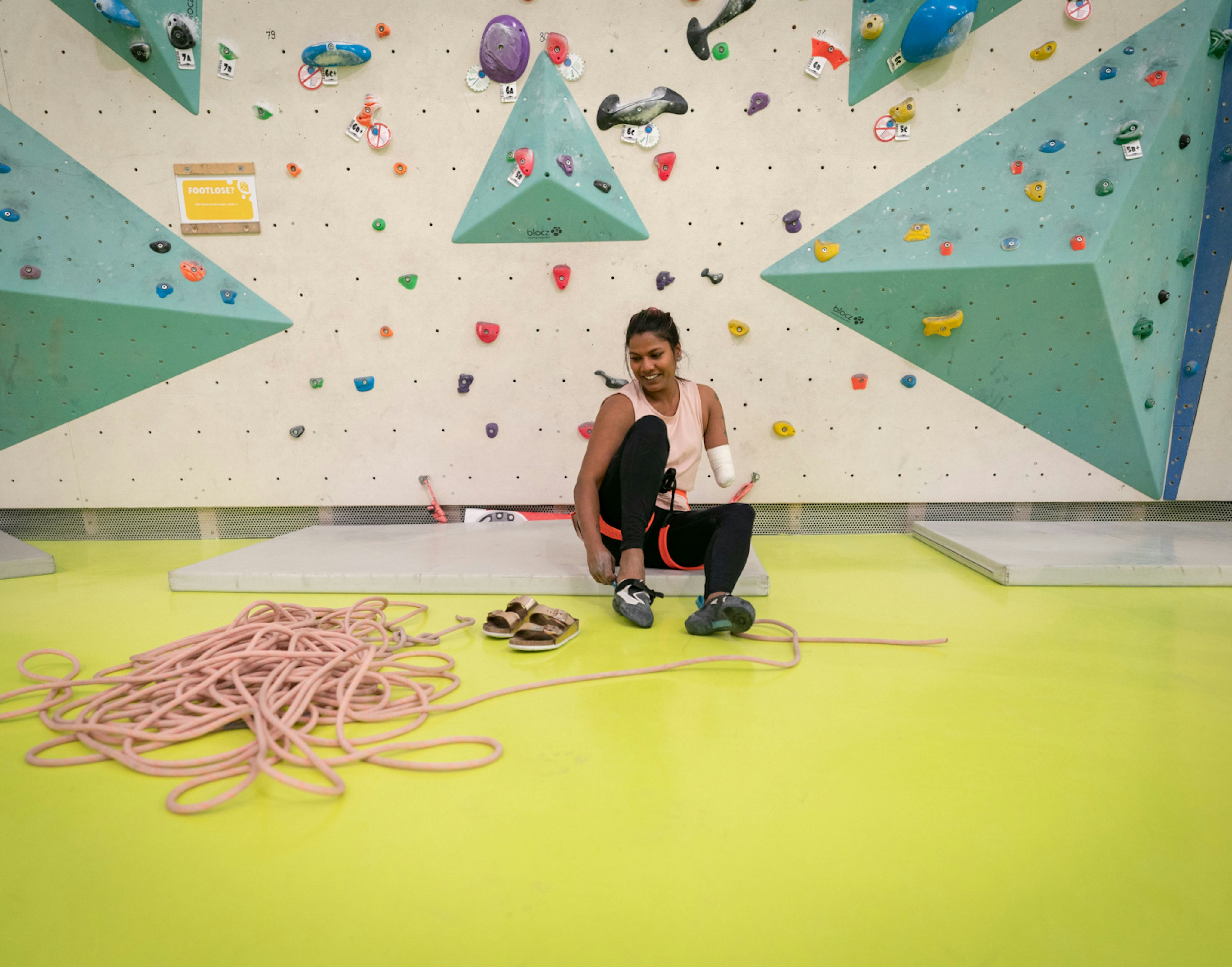 A woman sitting on the floor of a climbing gym, preparing for a climb by putting on Mammut climbing shoes next to a coil of Mammut rope.