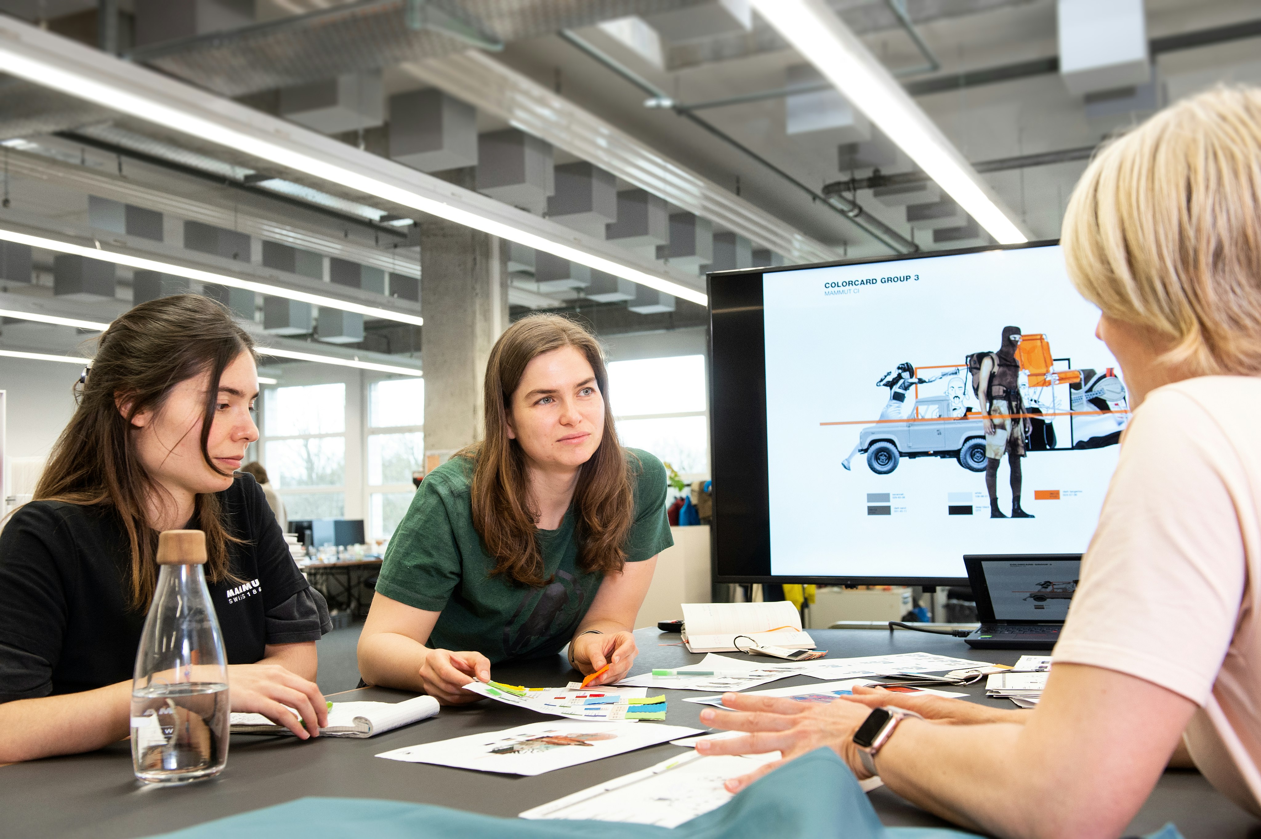 Three women, likely Mammut designers, discuss detailed design concepts around a table; a screen in the background displays an outdoor gear design.