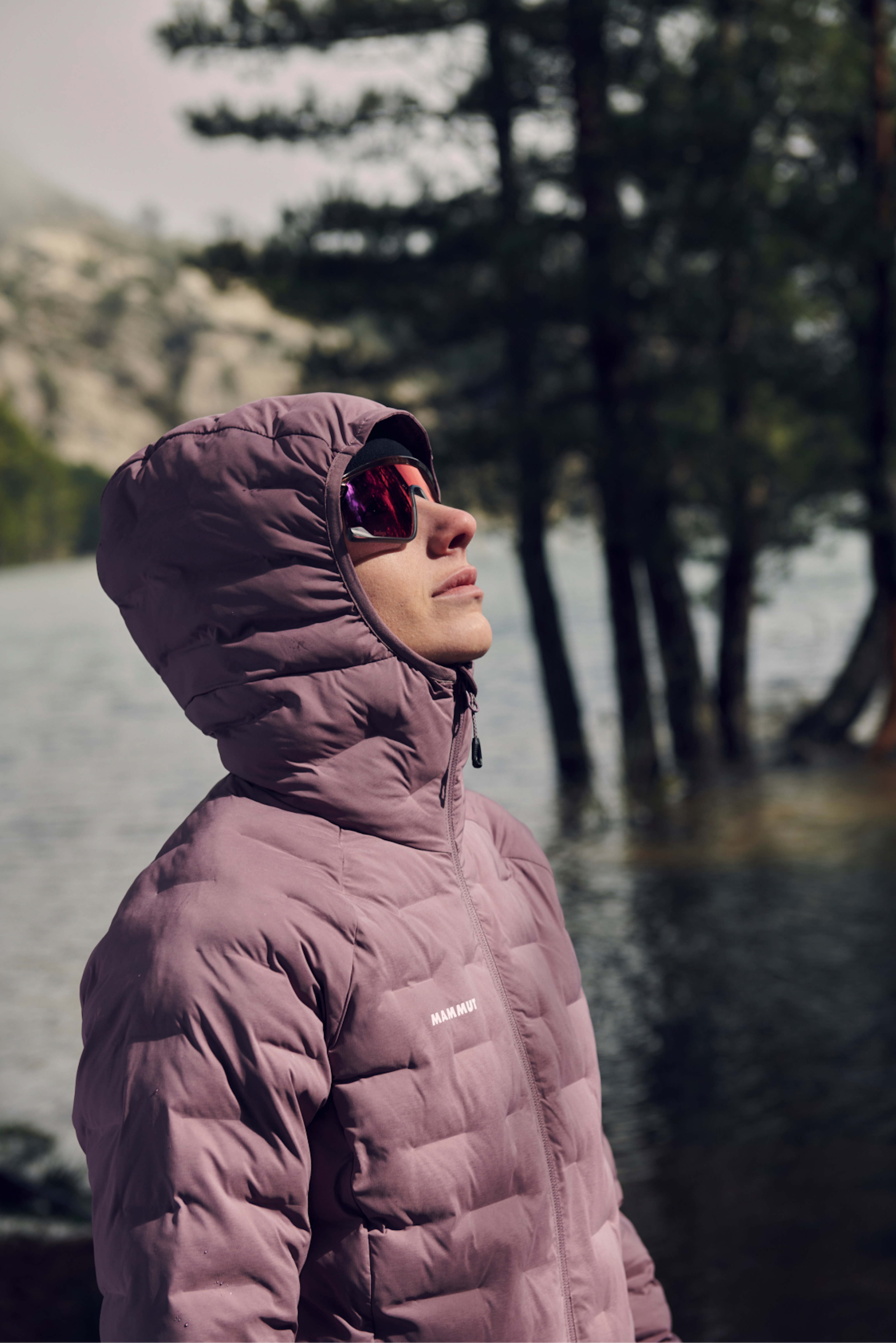 Mammut outdoor enthusiast in a hooded jacket and sunglasses, gazing up beside a serene lake surrounded by lush trees.