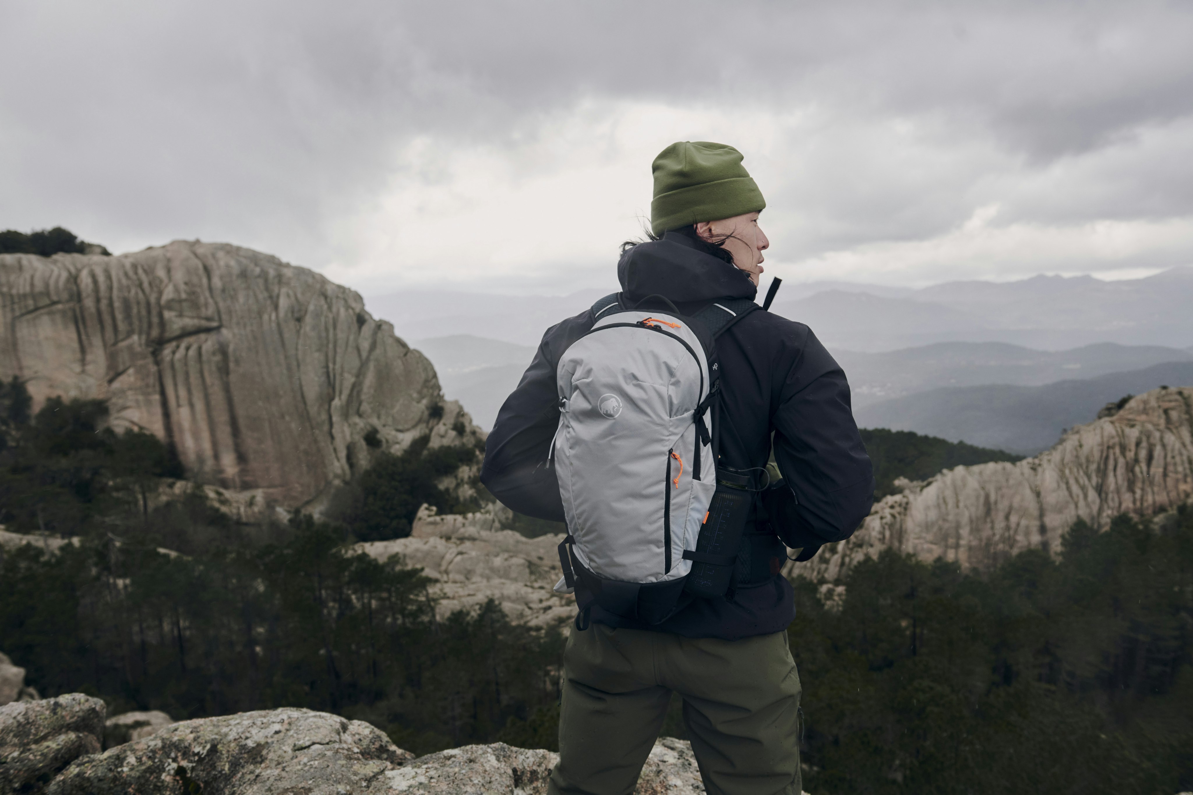 Person wearing a green Mammut hat and backpack standing on a rocky mountain terrain with cloudy skies in the background.