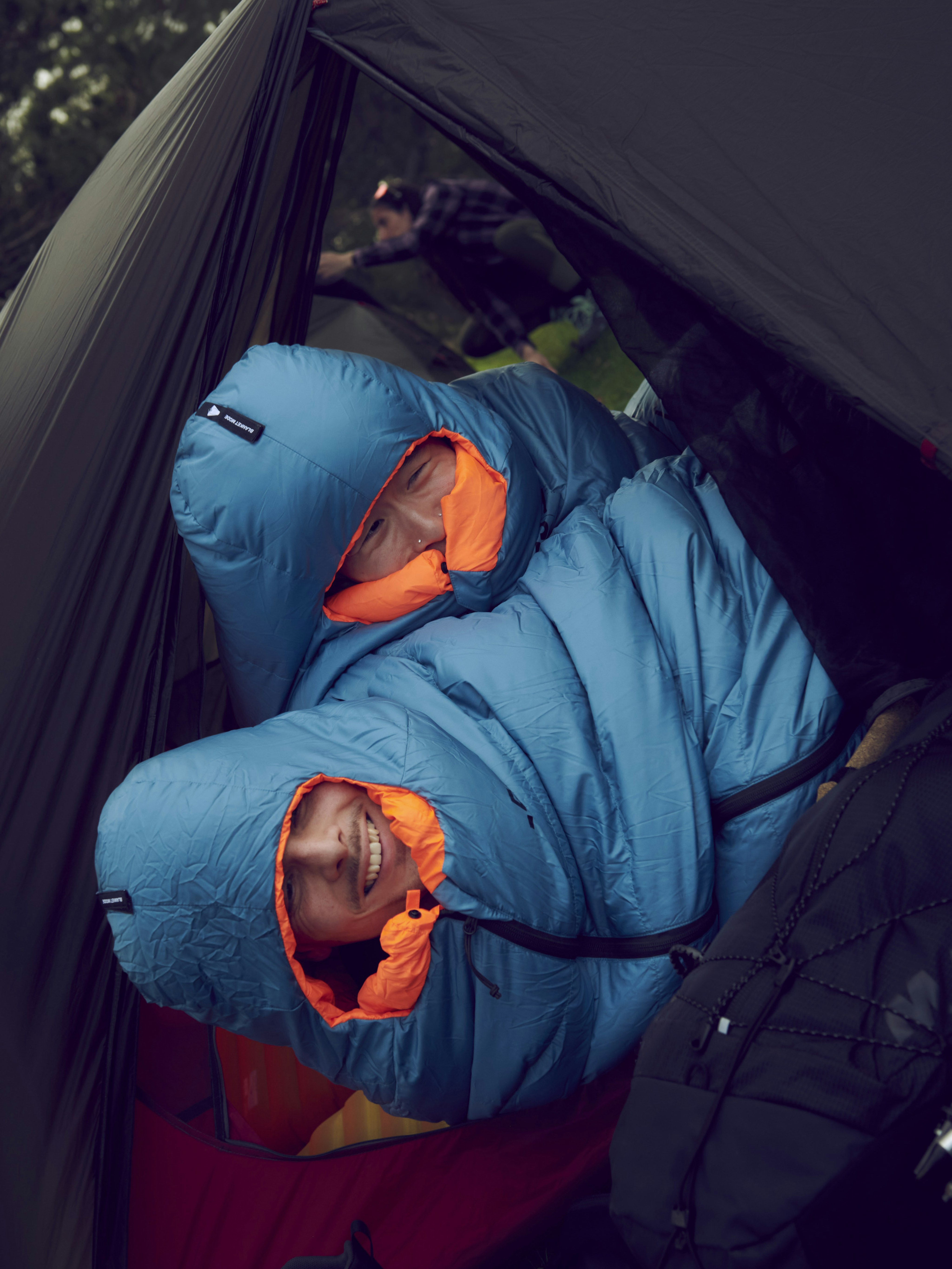 Two adventurers in Mammut blue sleeping bags, smiling inside a tent; another camper in the background outside the tent.