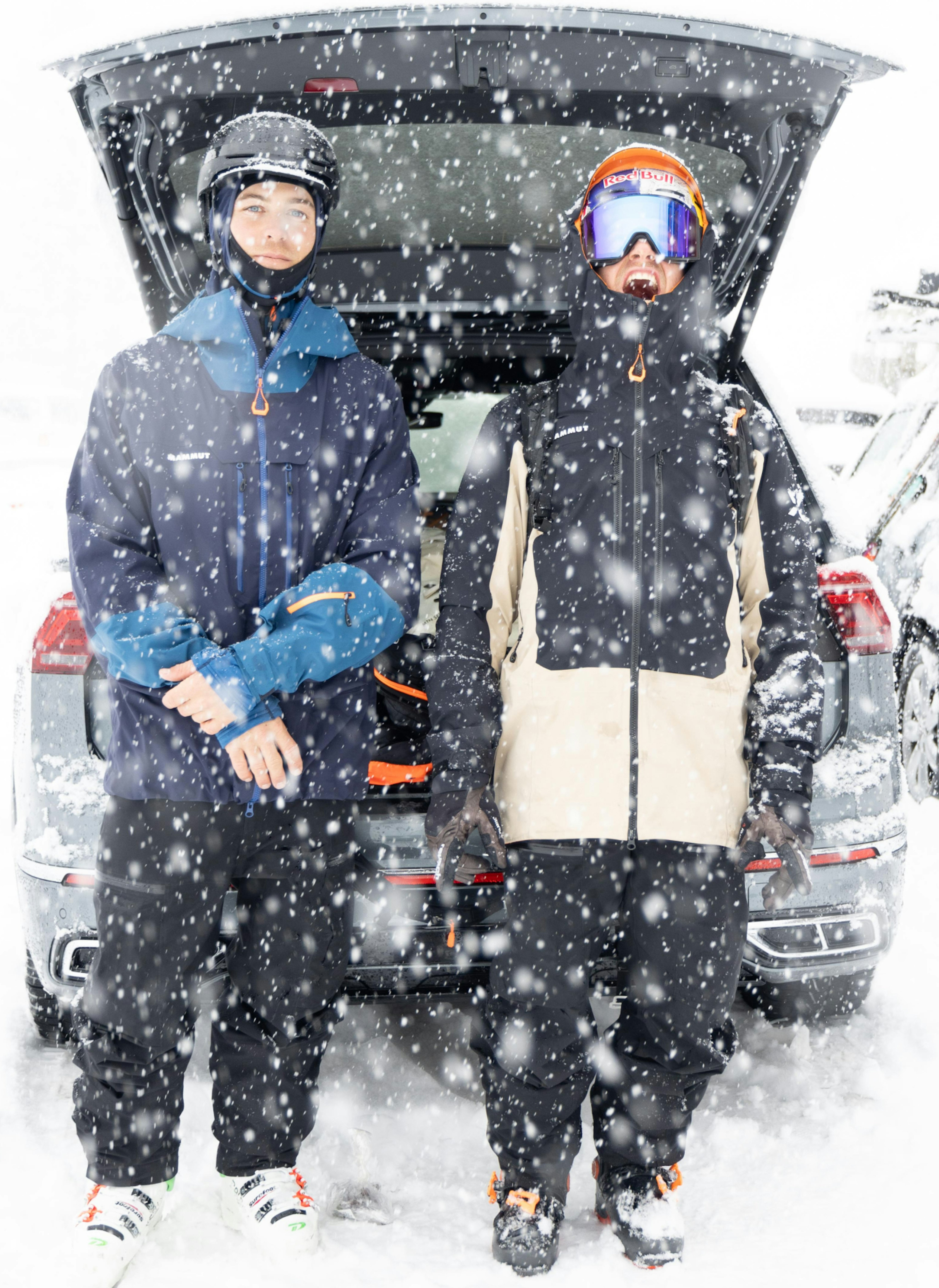 Two people in Mammut winter gear stand by an open car trunk, prepared for a snowy mountaineering adventure amidst falling snow.