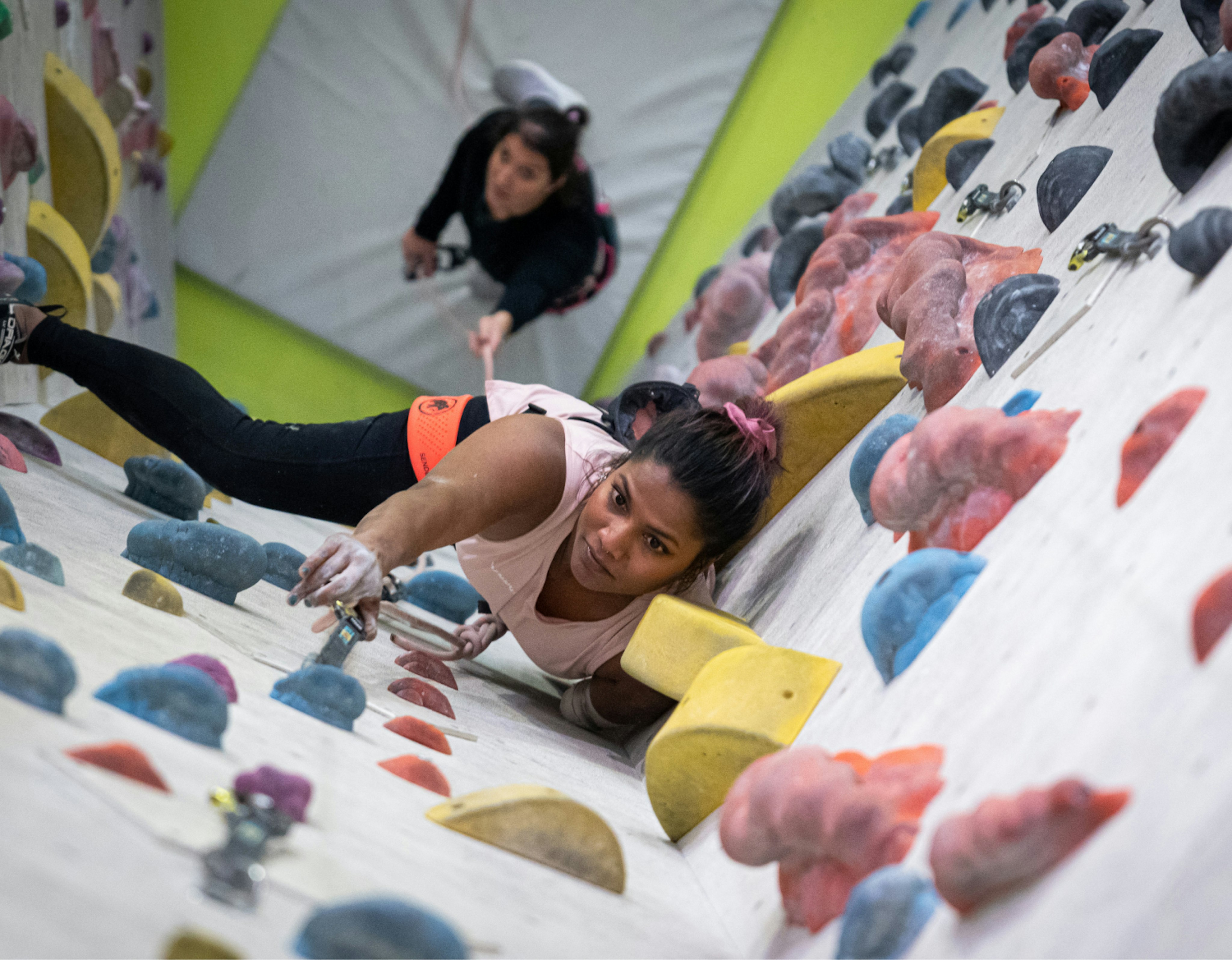 Two women rock climbing indoors at a Mammut-sponsored facility, focusing intensely while gripping colorful holds on an angled climbing wall.