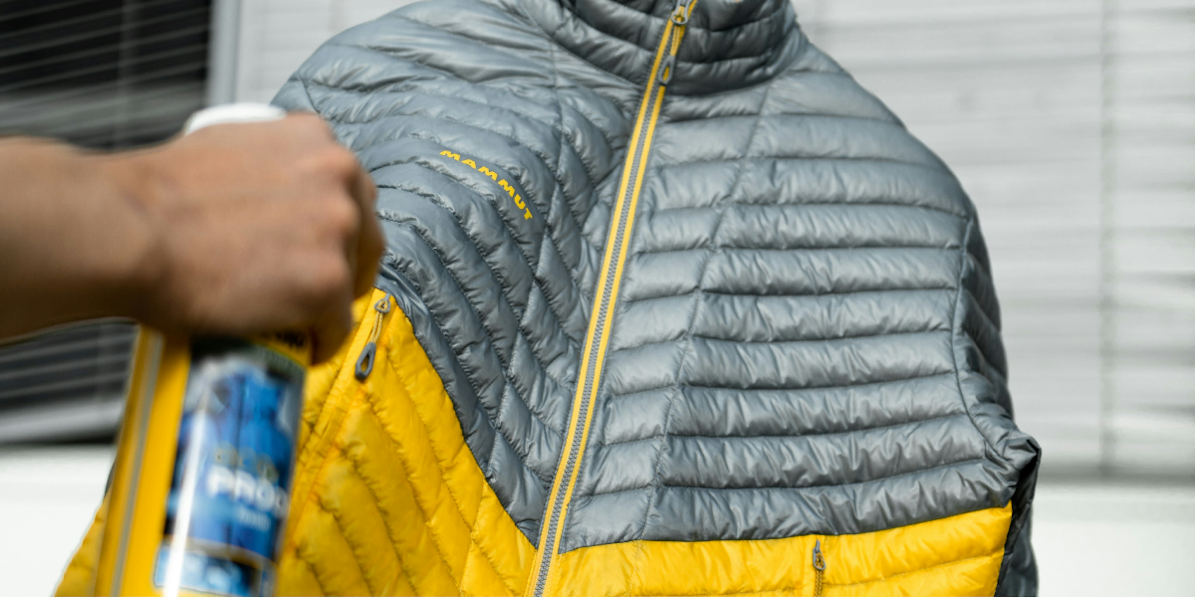 A technician spray coating a gray and yellow Mammut insulated jacket hanging on a rack, ensuring enhanced water resistance for outdoor adventures.
