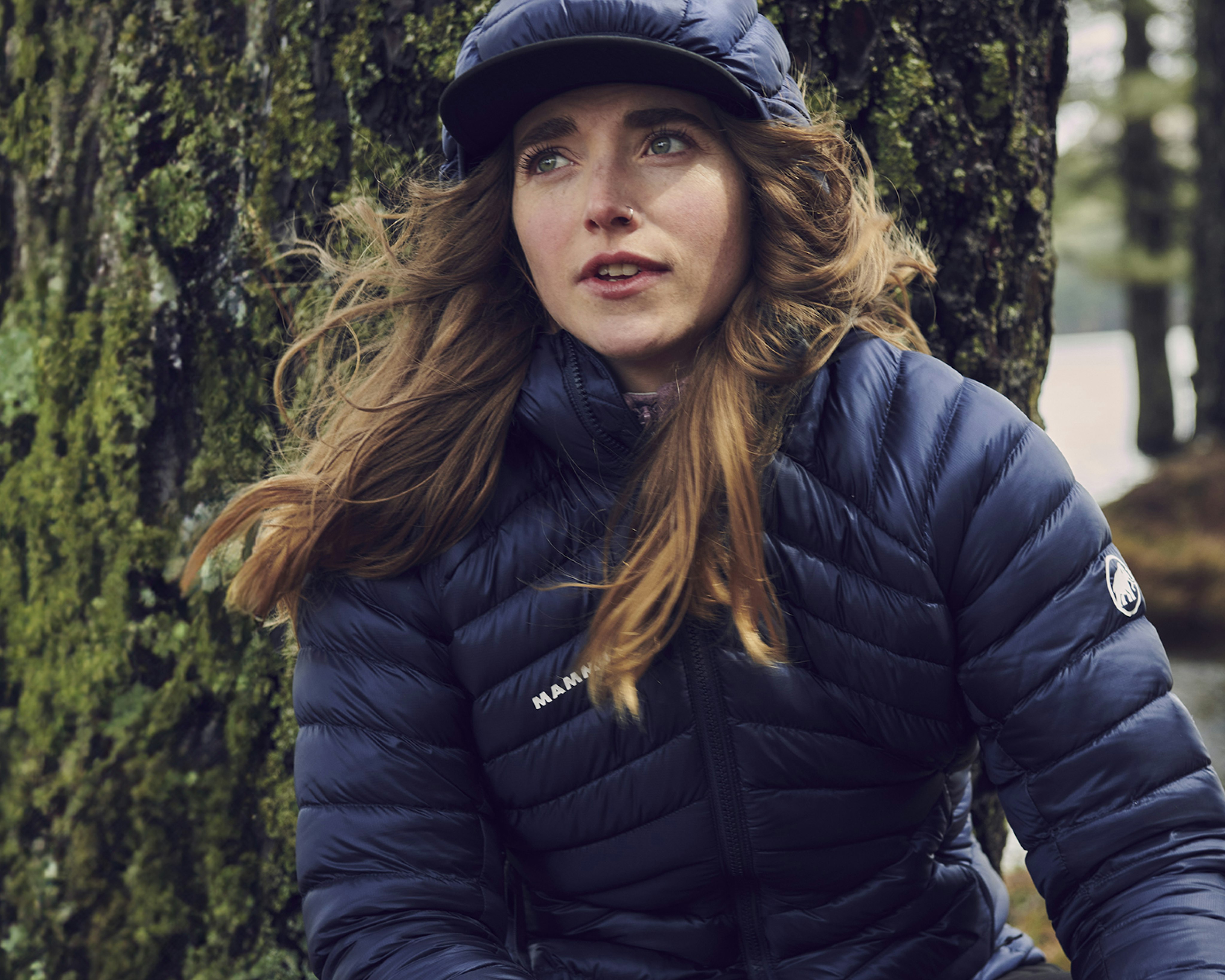 Woman in a blue Mammut puffy jacket and cap leans against a mossy tree in a lush forest setting.