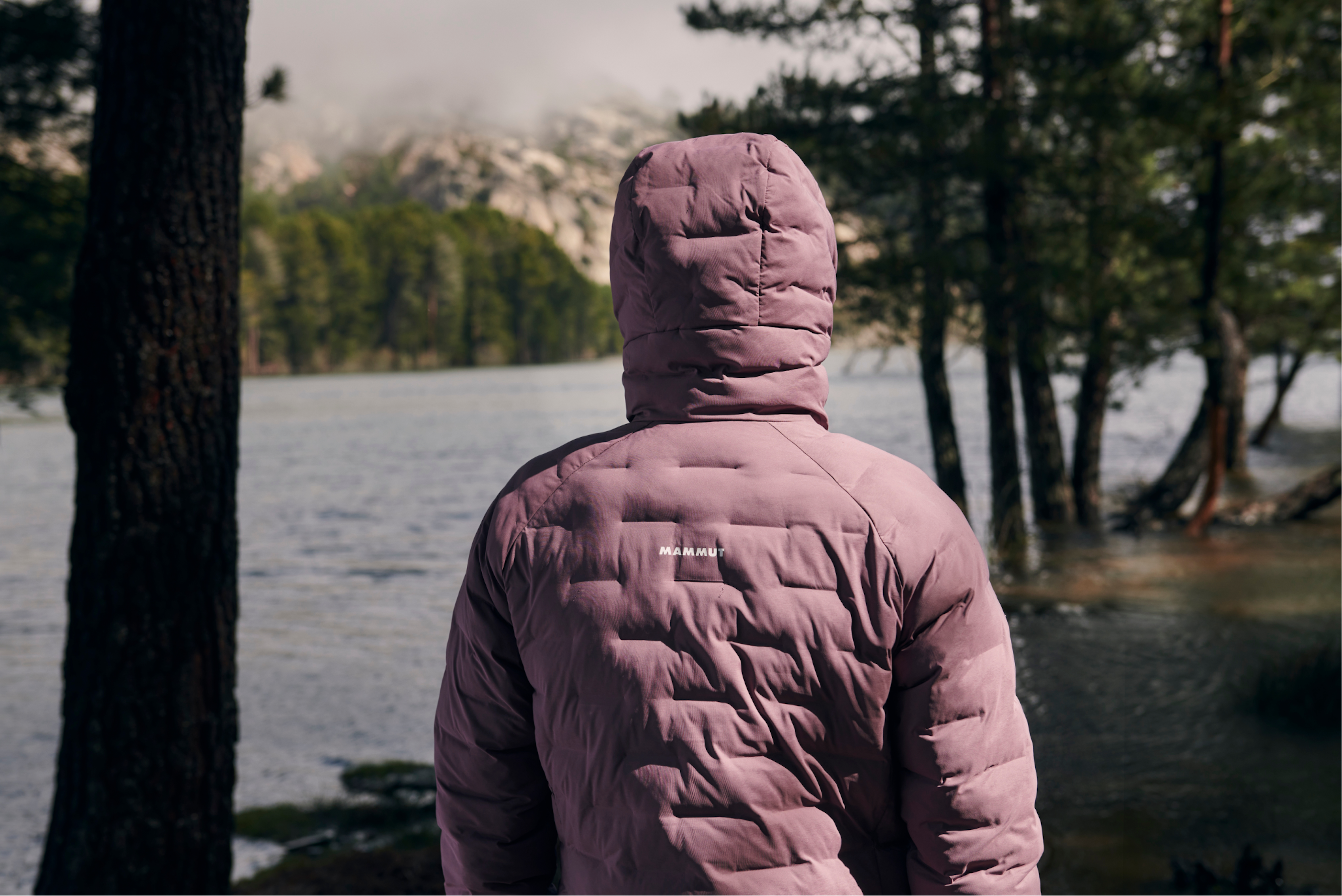 Mammut athlete in a pink puffer jacket standing near a foggy lake with tree-lined shores and towering mountains.