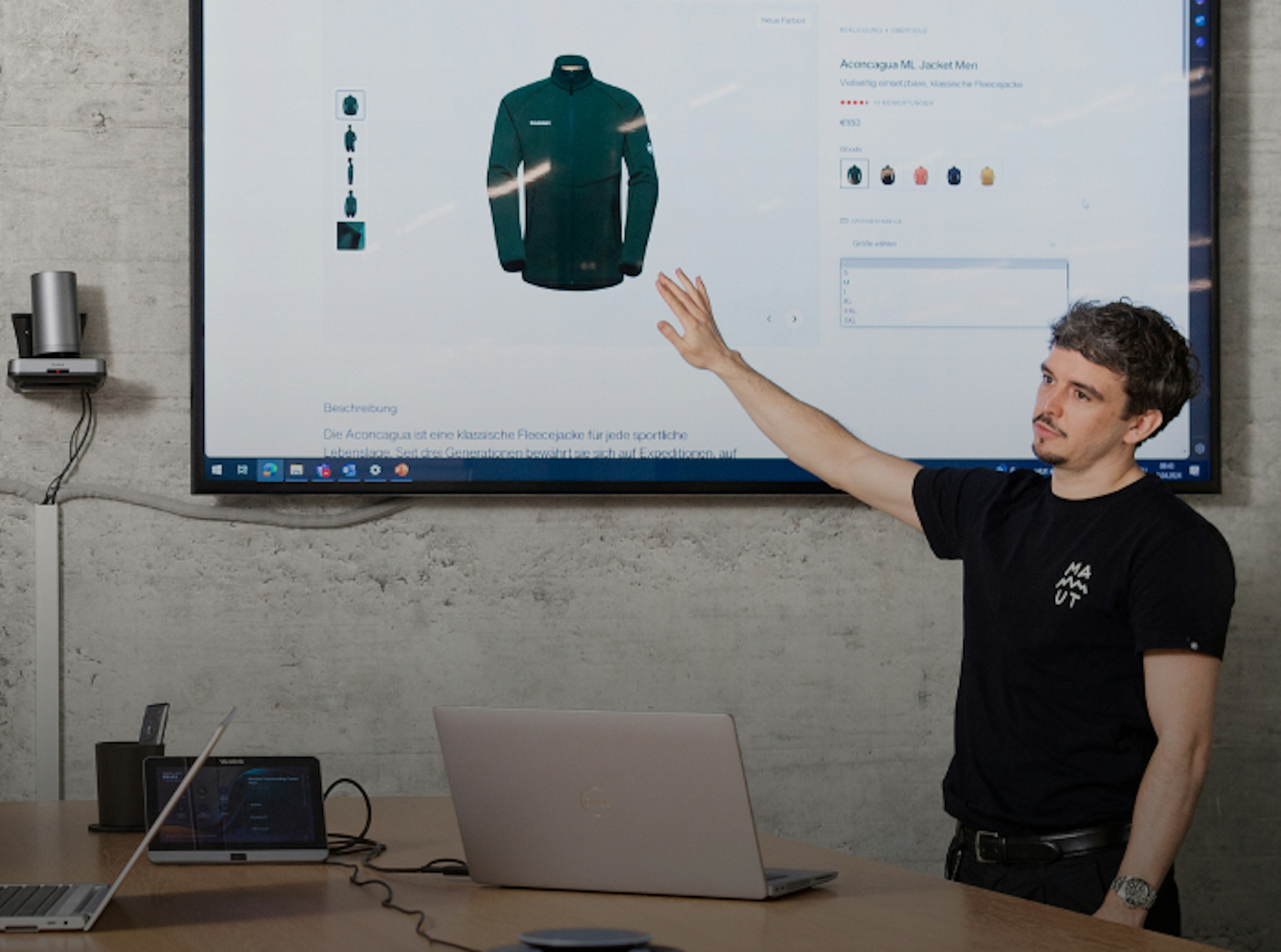 A man points to a large screen displaying a Mammut jacket product page during a presentation, with a laptop on the table.