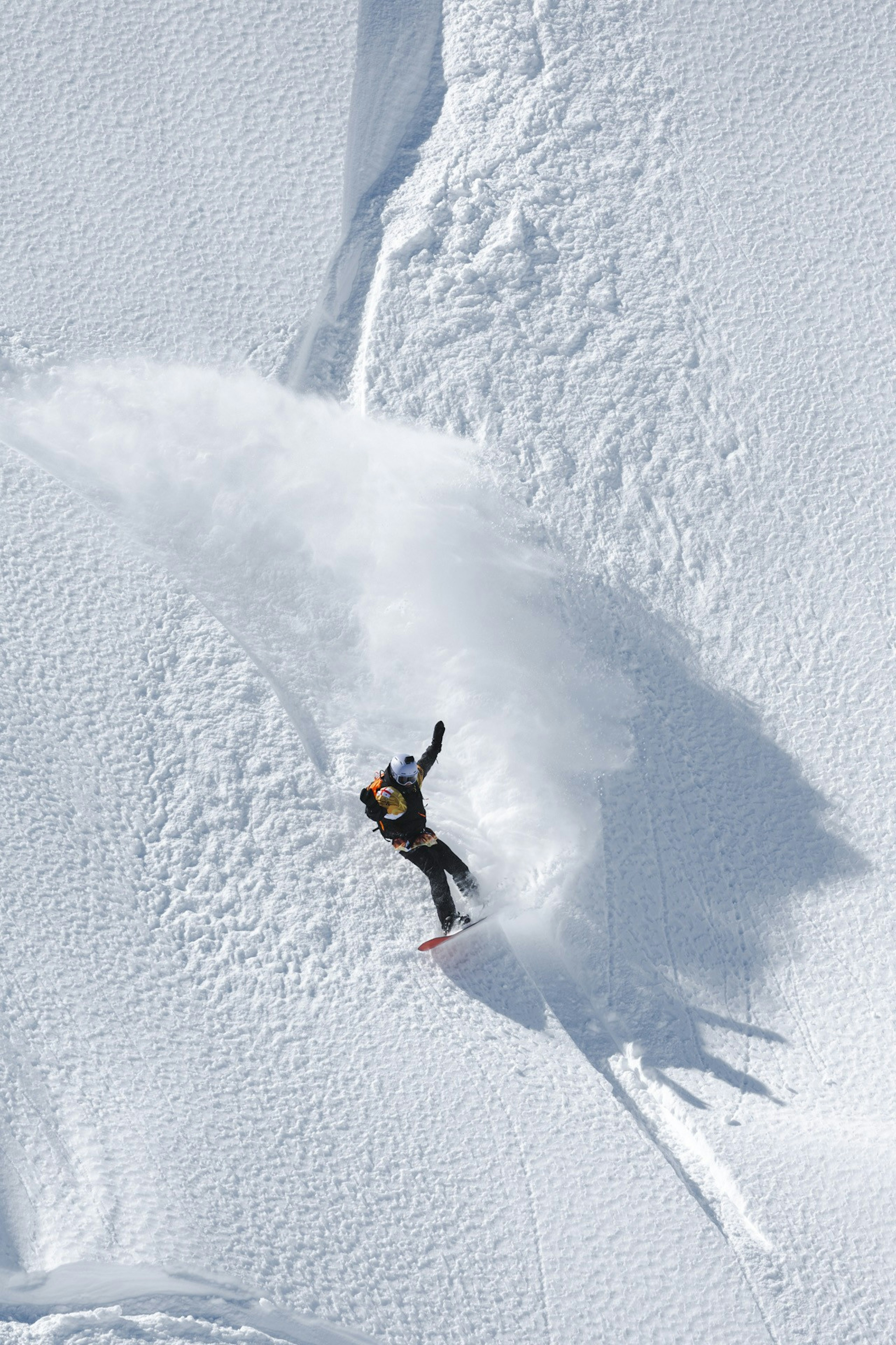 A snowboarder carves through fresh powder on a steep, snowy slope, creating a trail of snow spray behind them, showcasing Mammut's high-performance winter sports gear.