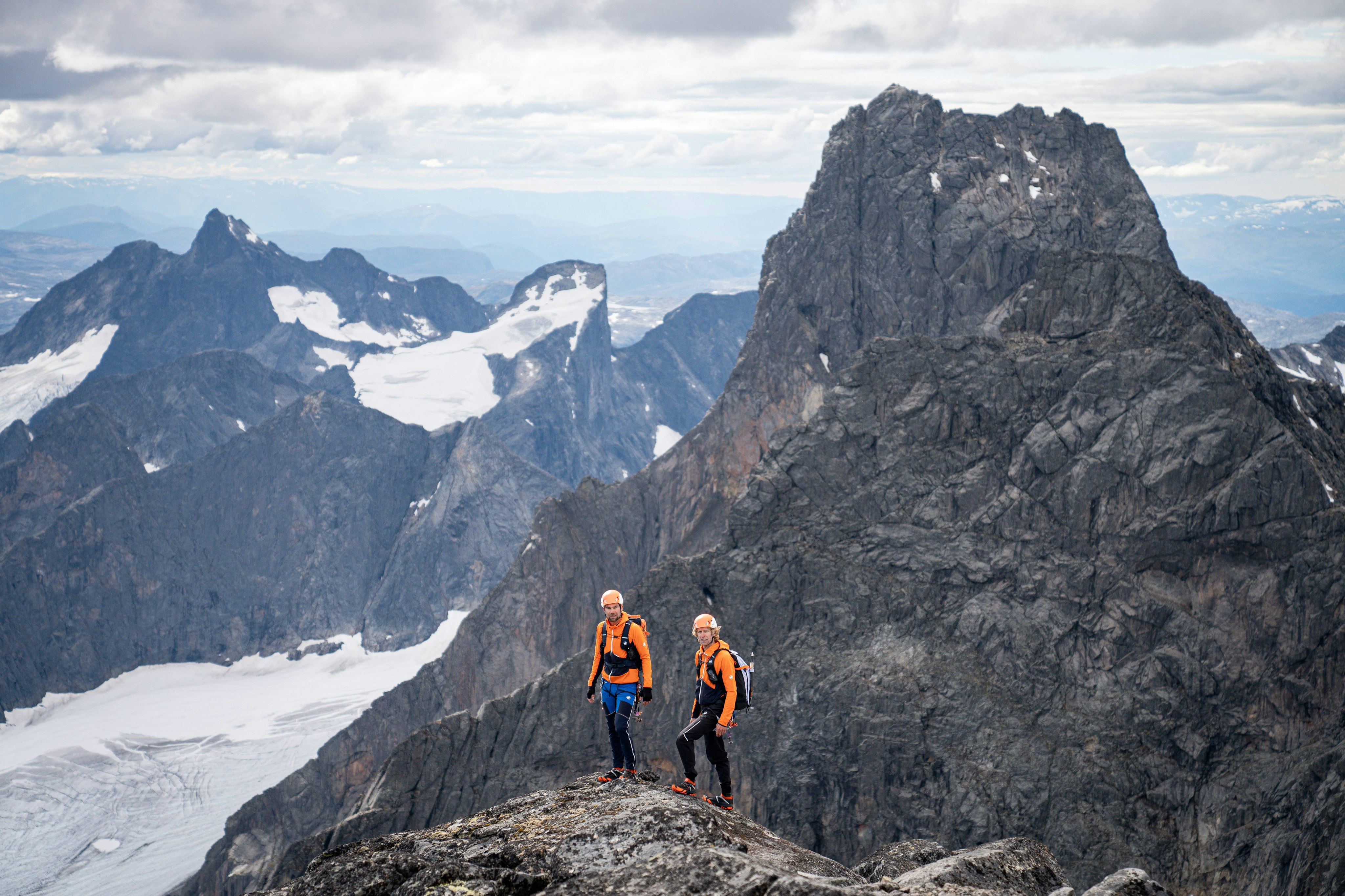 Two hikers wearing Mammut orange jackets standing on a rugged mountain peak with a vast alpine range and snow-capped peaks in the background.