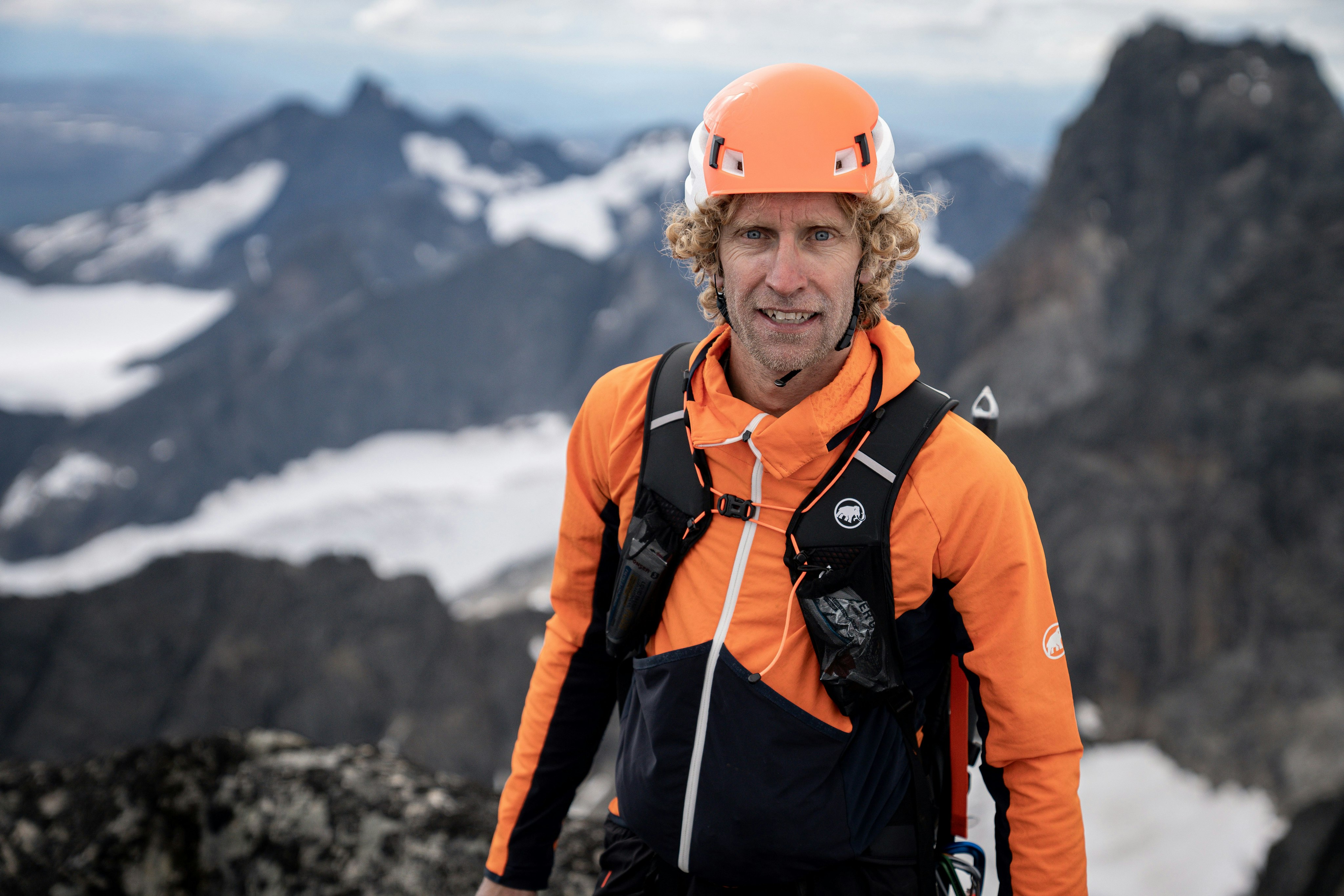 A climber in a Mammut orange helmet and jacket stands on a mountain surrounded by snowy peaks.
