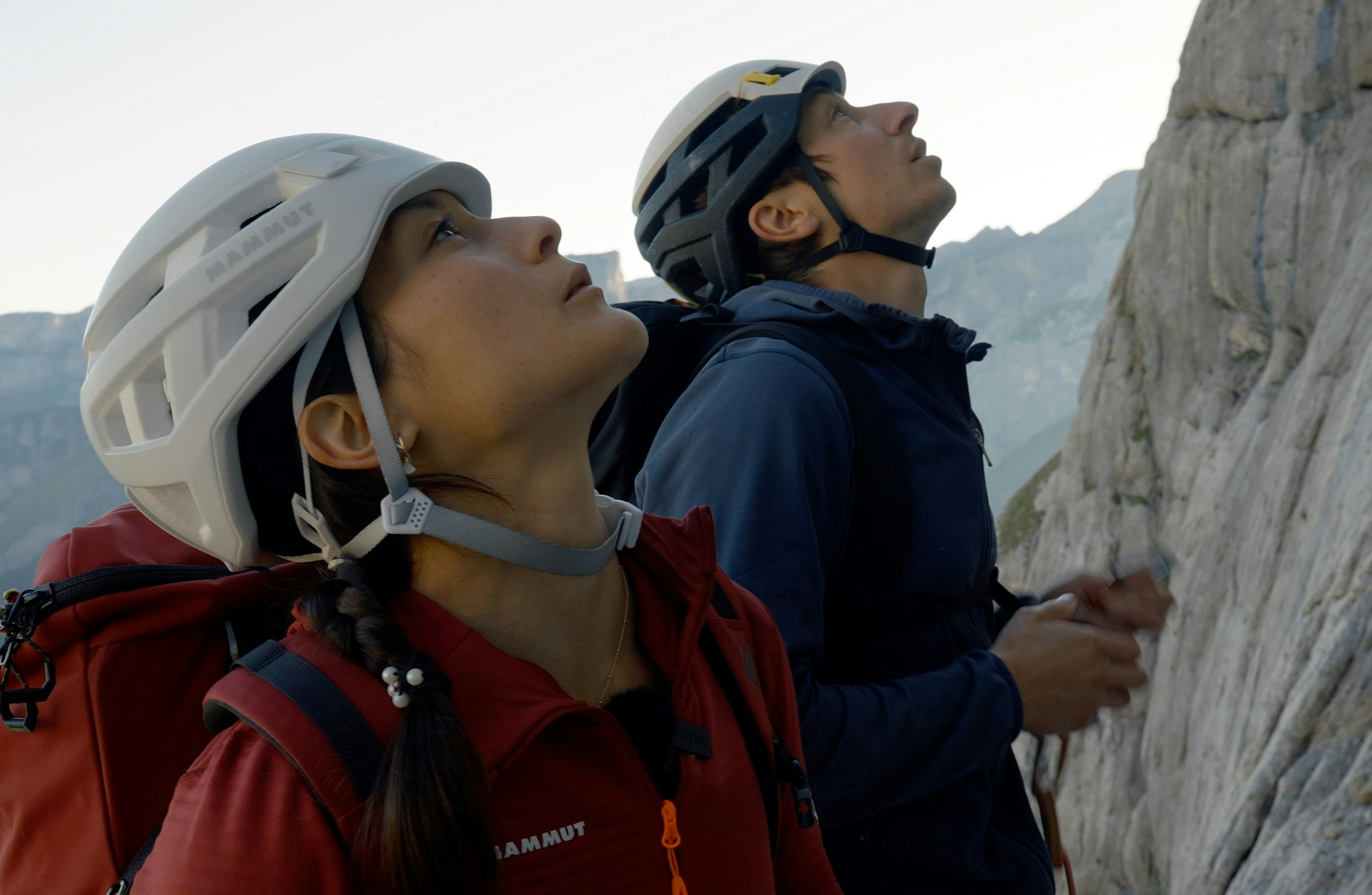 Katharine Choong, a climber wearing Mammut helmets and jackets look up while standing near a rocky cliff face.