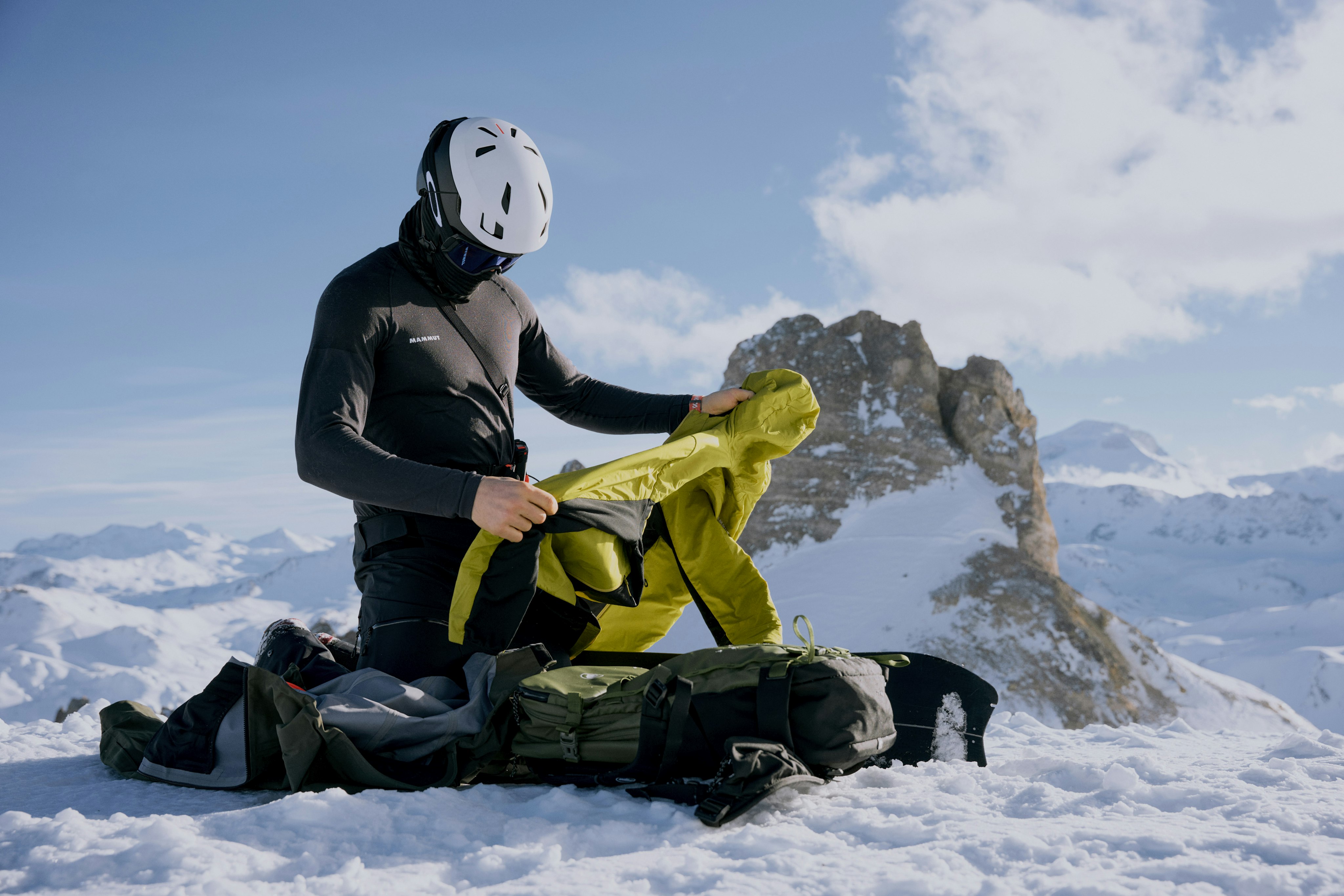 Two hikers wearing Mammut gear on a mountain trail, one looking back towards the camera. Snow-capped peaks are visible in the background.