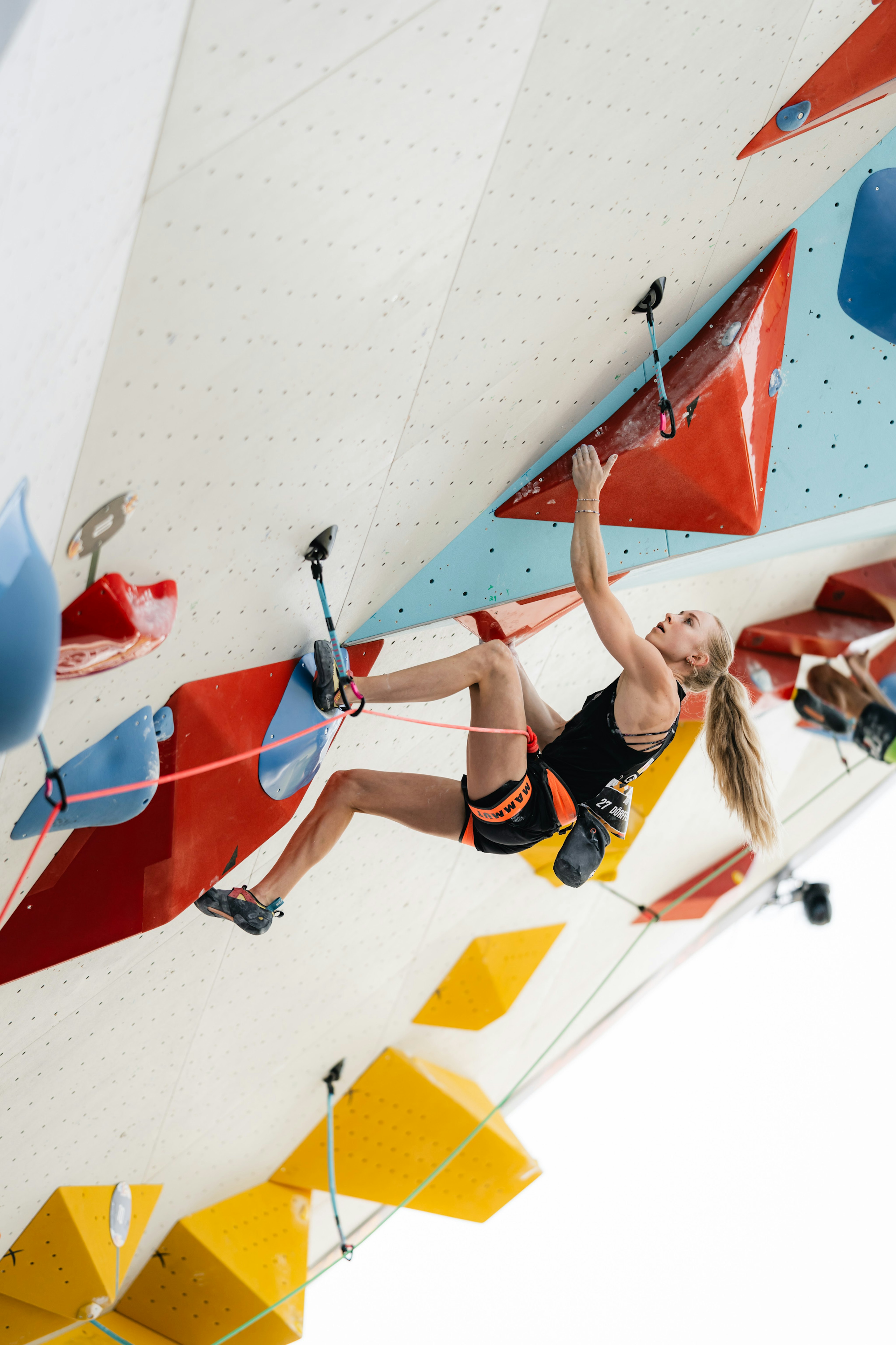 Lucia Dörffel a female climber in action on an indoor climbing wall featuring vibrant holds, showcasing her skills with Mammut gear against a stark white background.