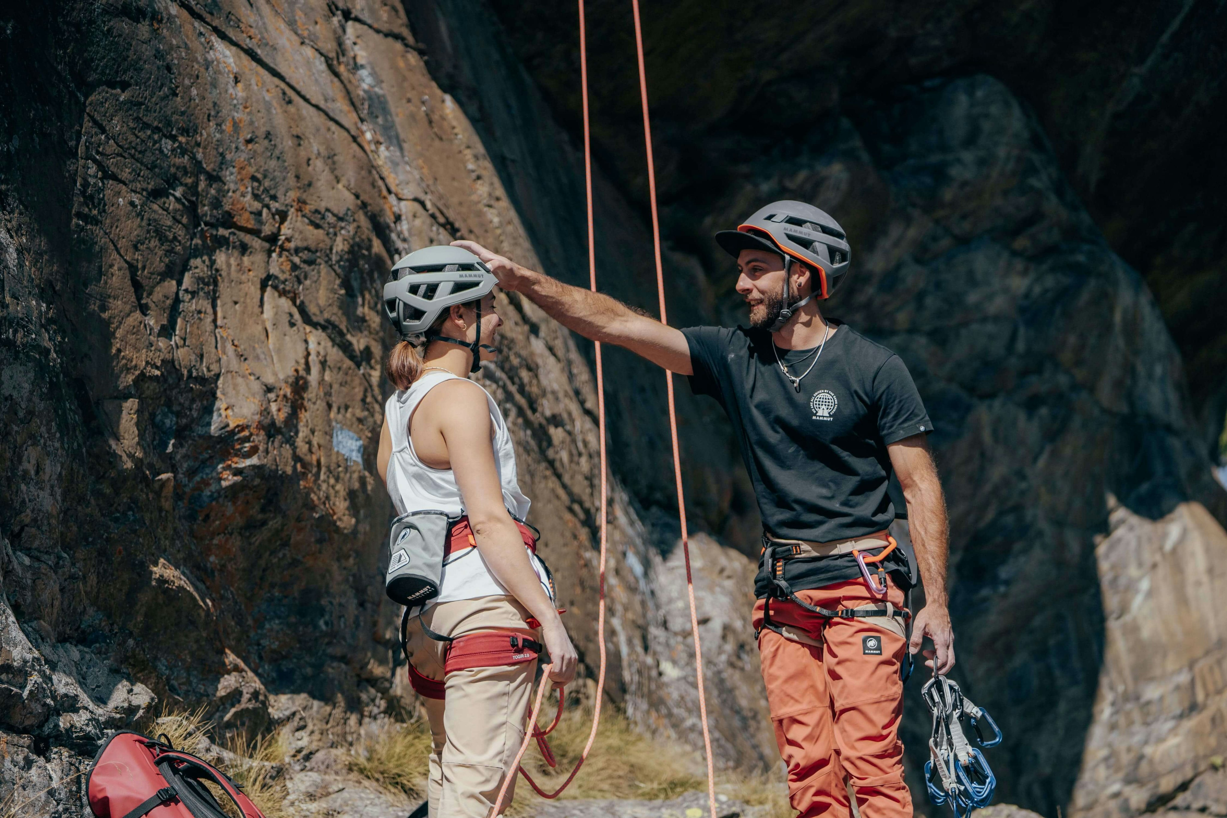 Two climbers equipped with Mammut helmets and climbing gear share a light-hearted moment at the base of a rugged rocky cliff.