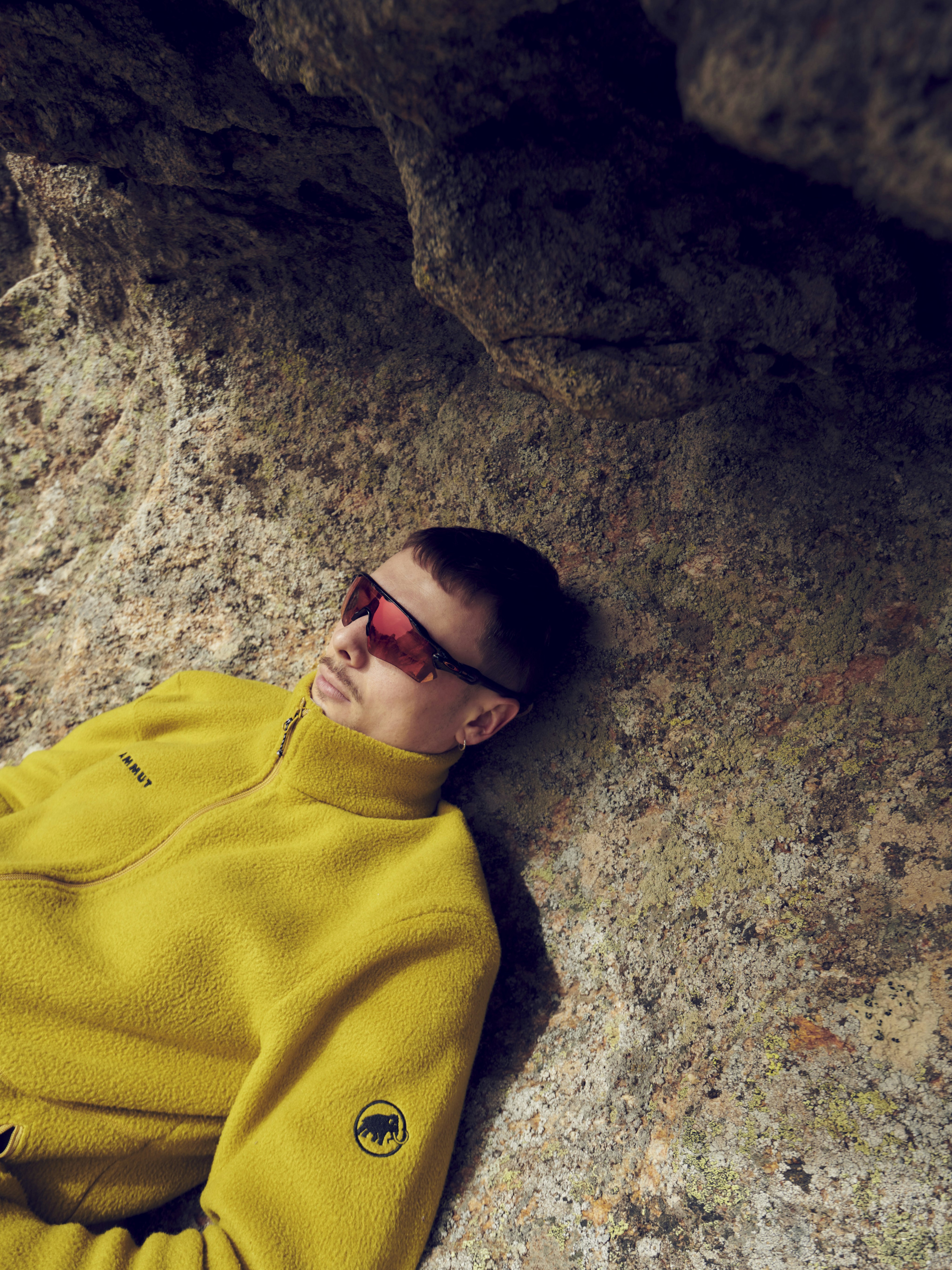 Person in a yellow Mammut jacket and red sunglasses leans against a rocky surface, looking relaxed and contemplative.