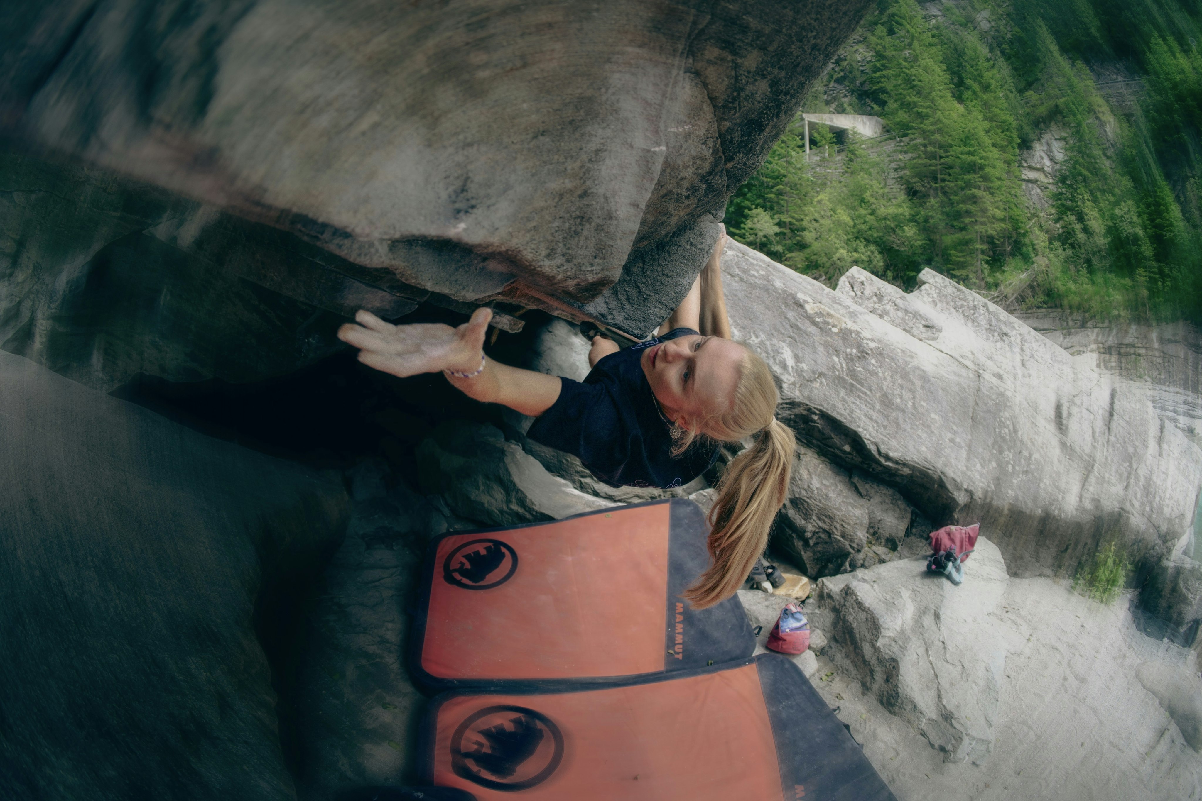 Rock climber Lucia Dörffel equipped with Mammut gear, reaching up with one hand while surrounded by rocky terrain and greenery, with a safety mat below.