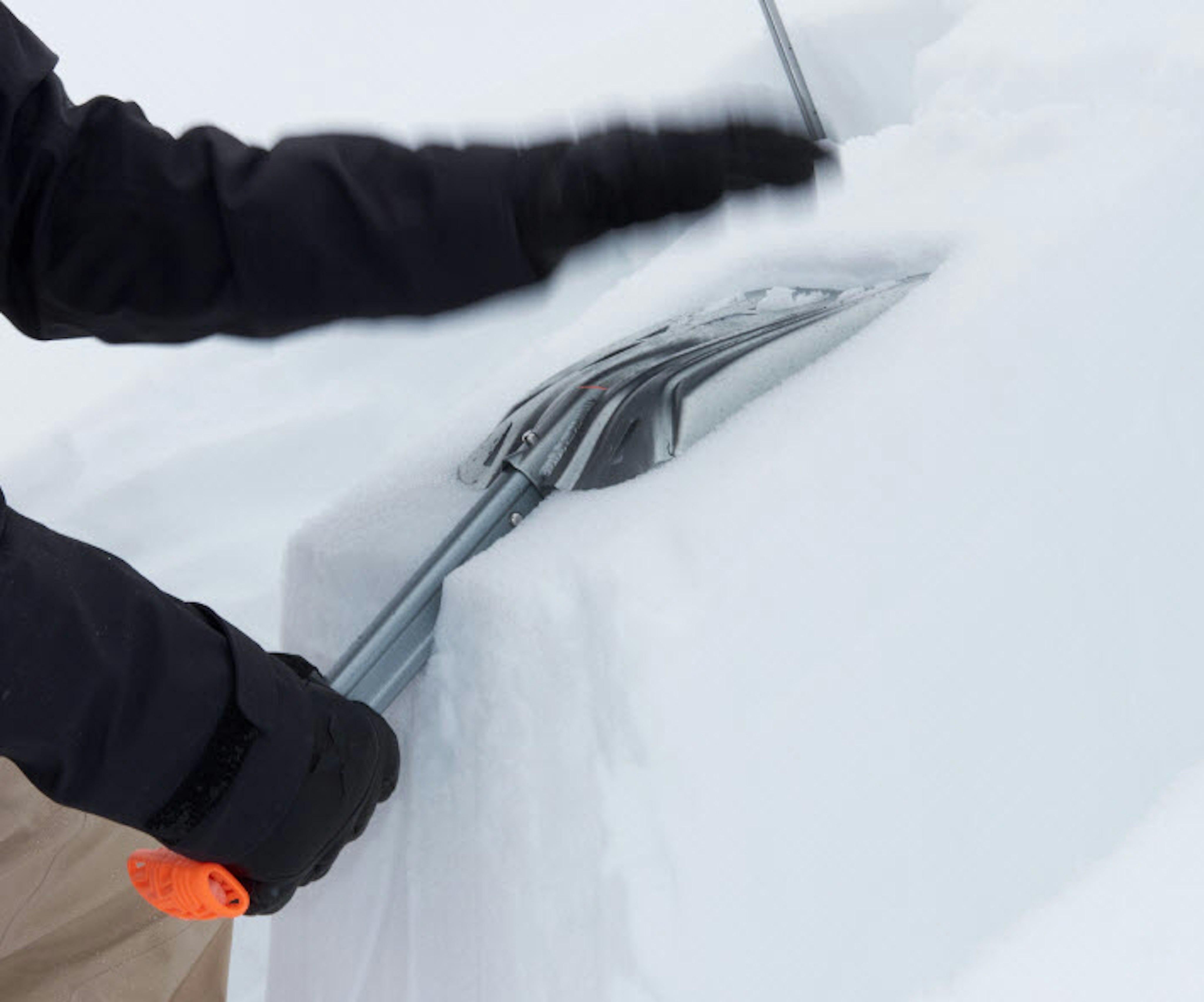 A person clearing a thick layer of snow with a plastic snow shovel on a snowy day, wearing Mammut outdoor gear.