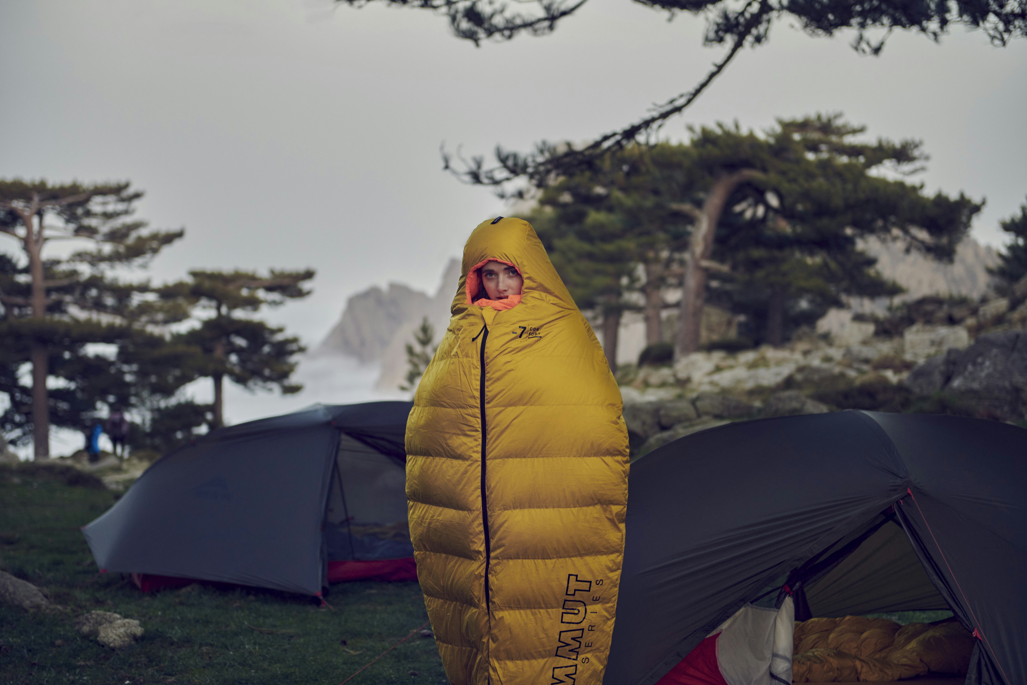 Explorer wearing a yellow Mammut sleeping bag in front of tents and trees, embracing the adventure outdoors.