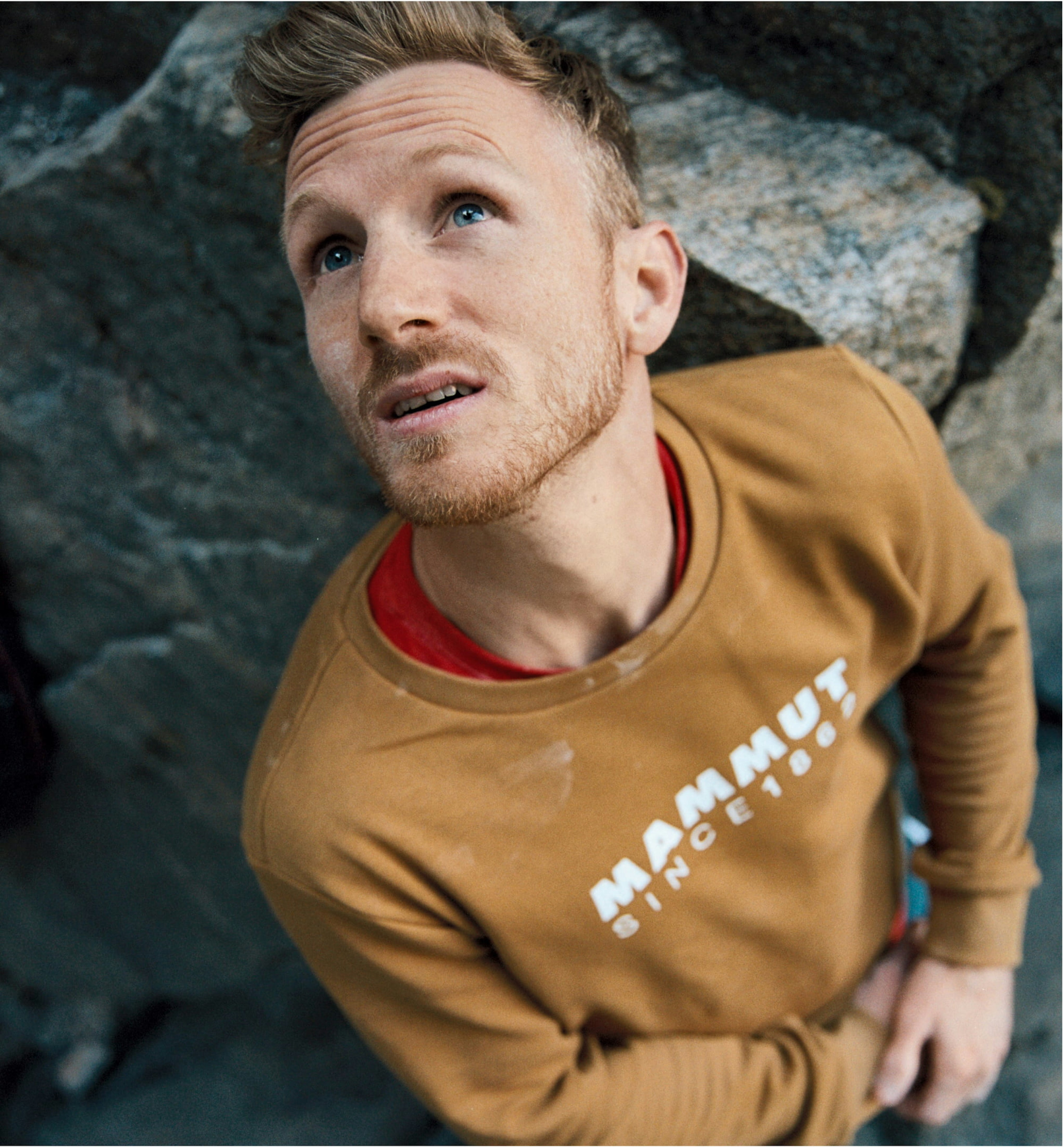 A man wearing a tan Mammut sweatshirt gazes thoughtfully upwards against a rugged rock background.