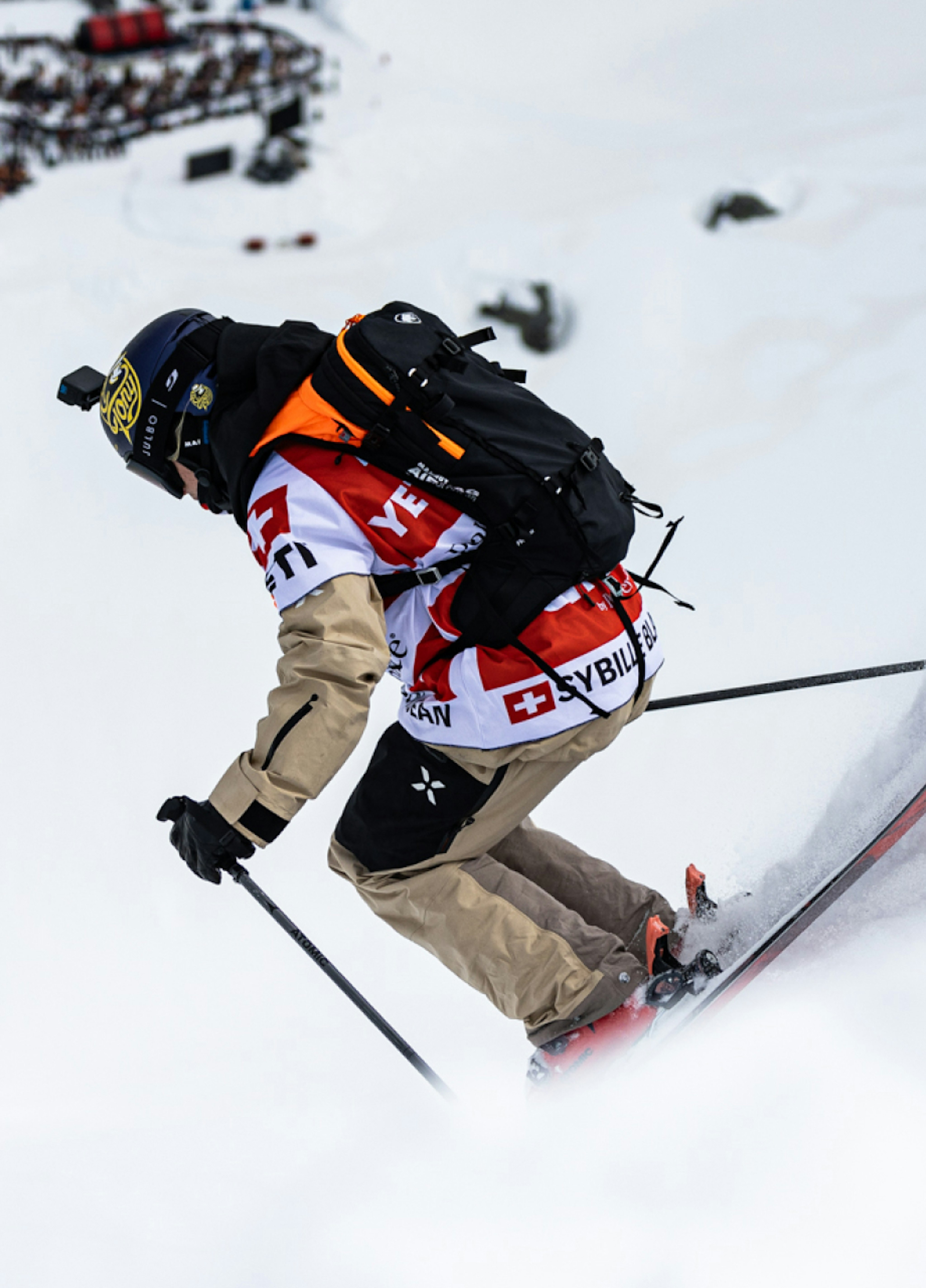 A skier in a beige Mammut outfit and black helmet descends a snowy slope during a race, displaying various sponsor logos.