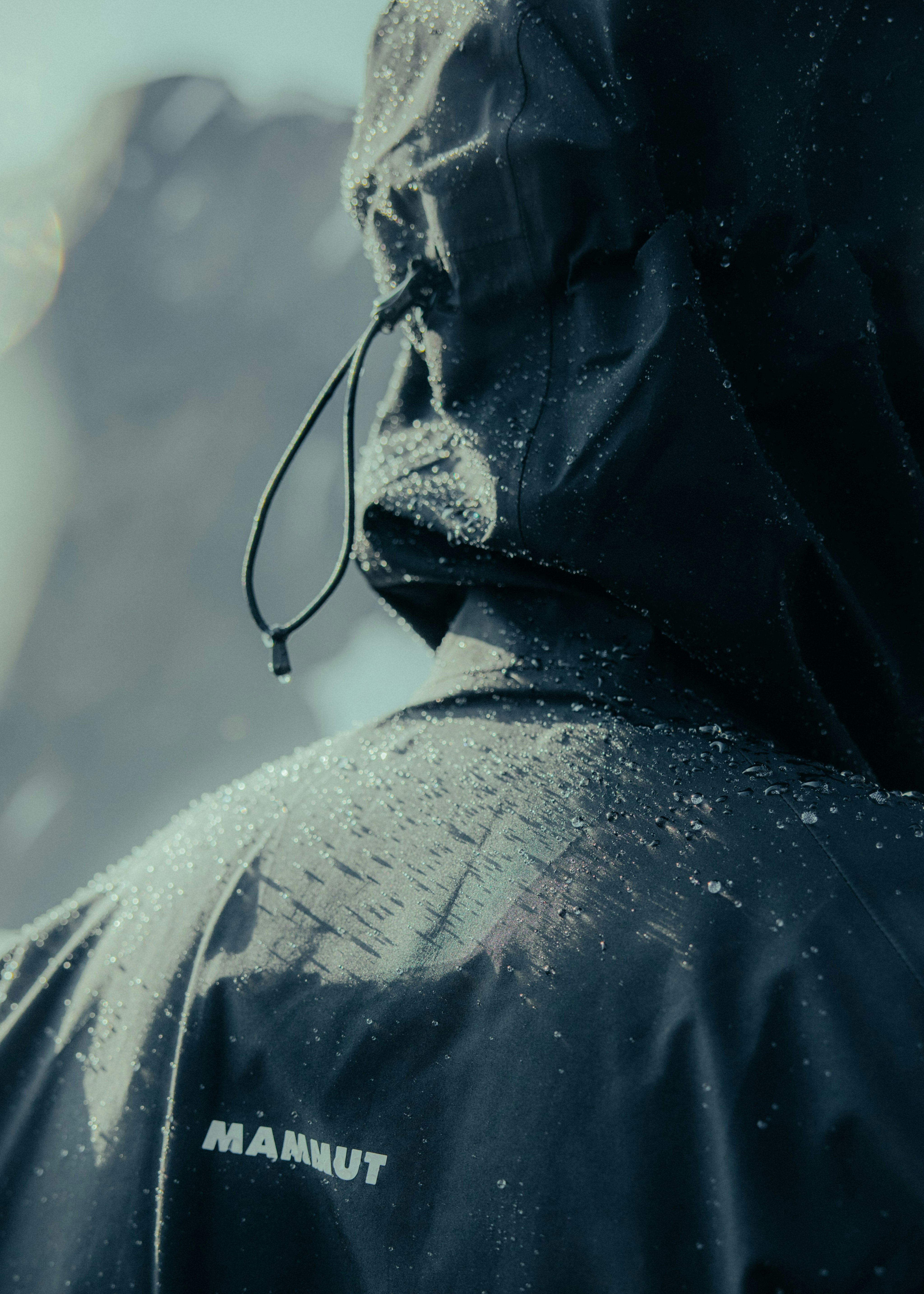 A person in a black Mammut waterproof jacket with a hood, covered in droplets of water, stands facing away from the camera amidst a foggy mountain landscape.