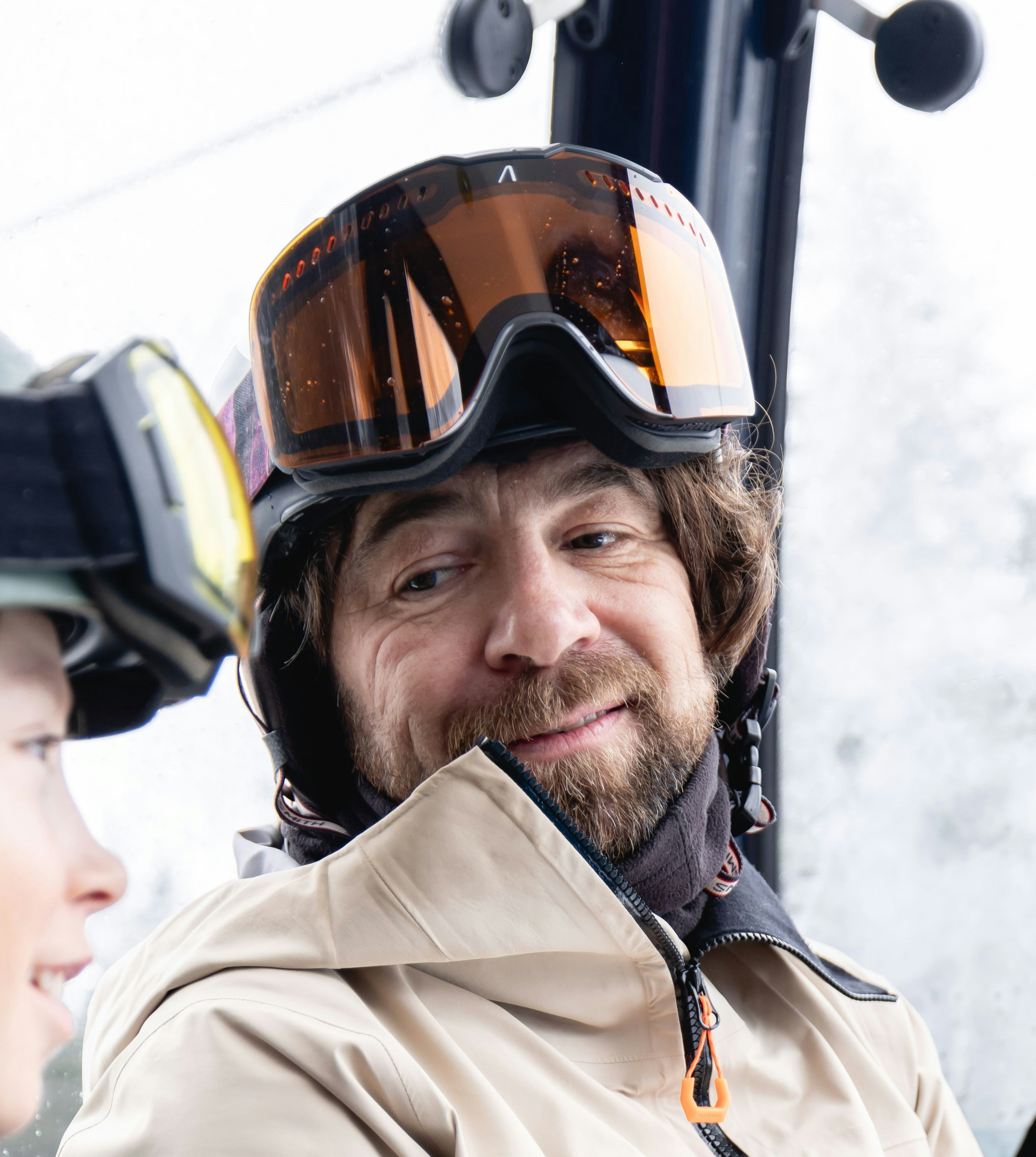 Whisky the tutor in Mammut ski gear with goggles smiles at a child inside a ski lift or gondola.