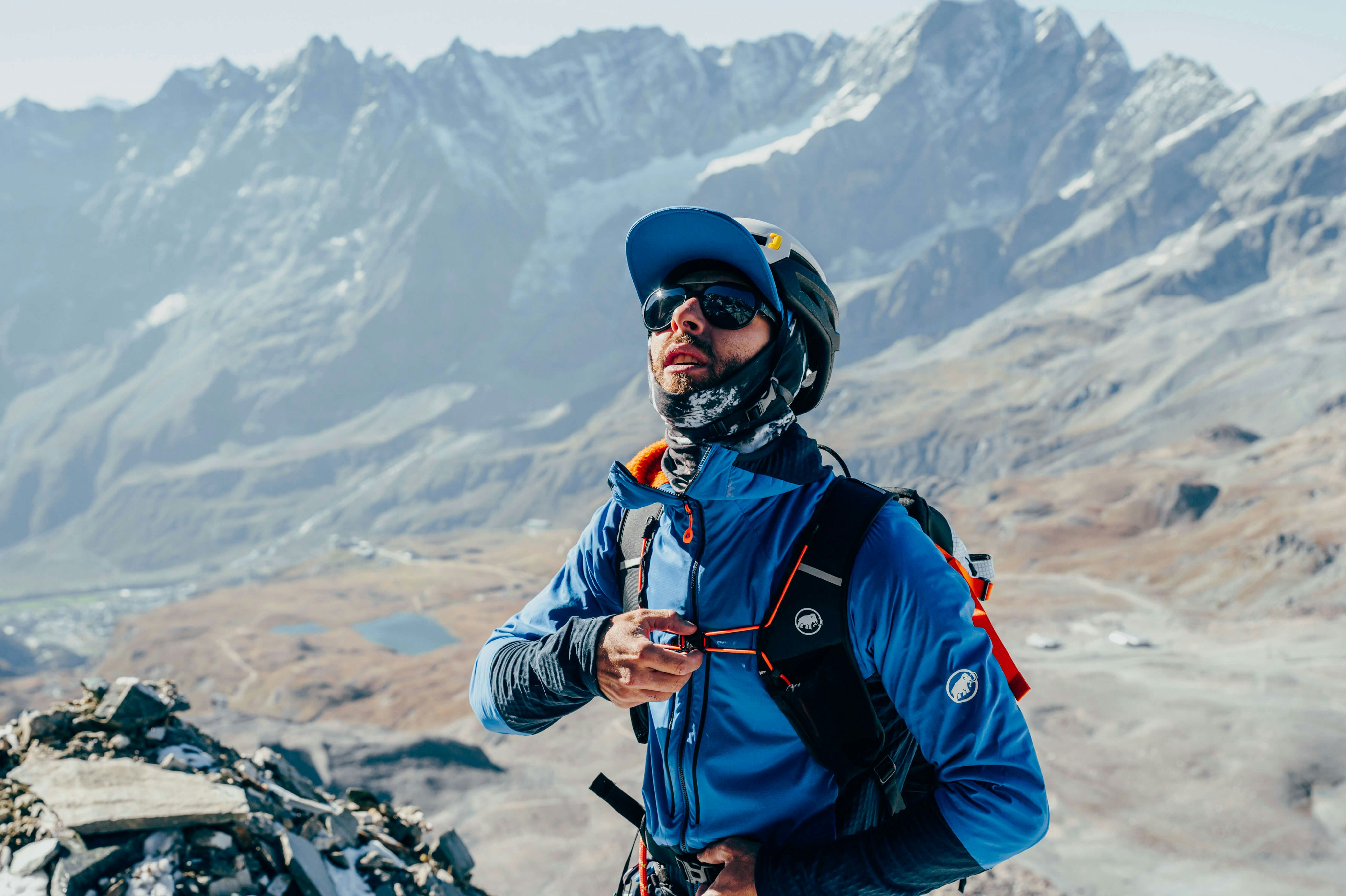 A mountaineer in blue Mammut gear and a helmet stands on a rocky summit with snowy mountains in the background.