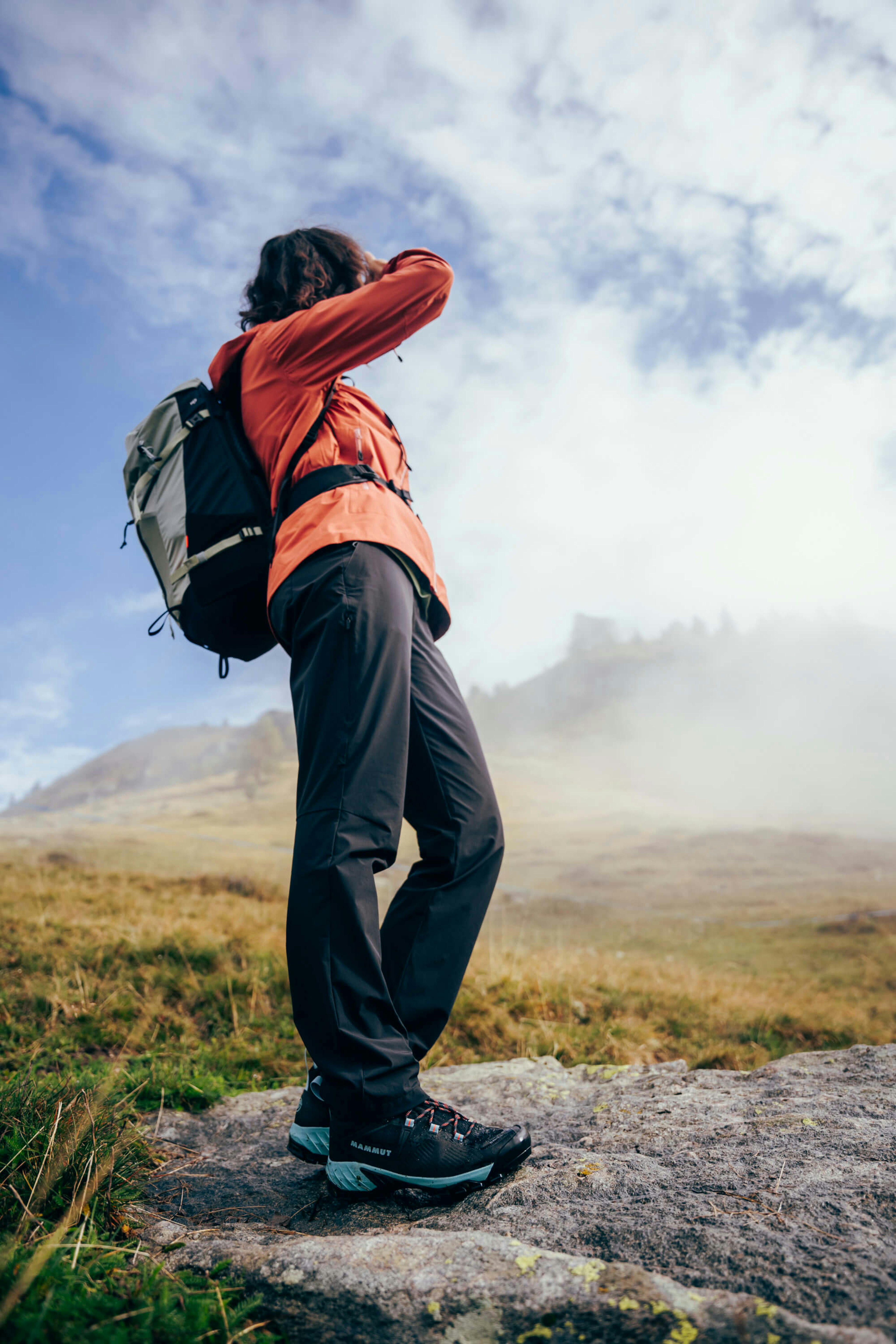 Person wearing a Mammut backpack and outdoor gear, standing on a rock, looking at the sky in a misty mountain landscape.