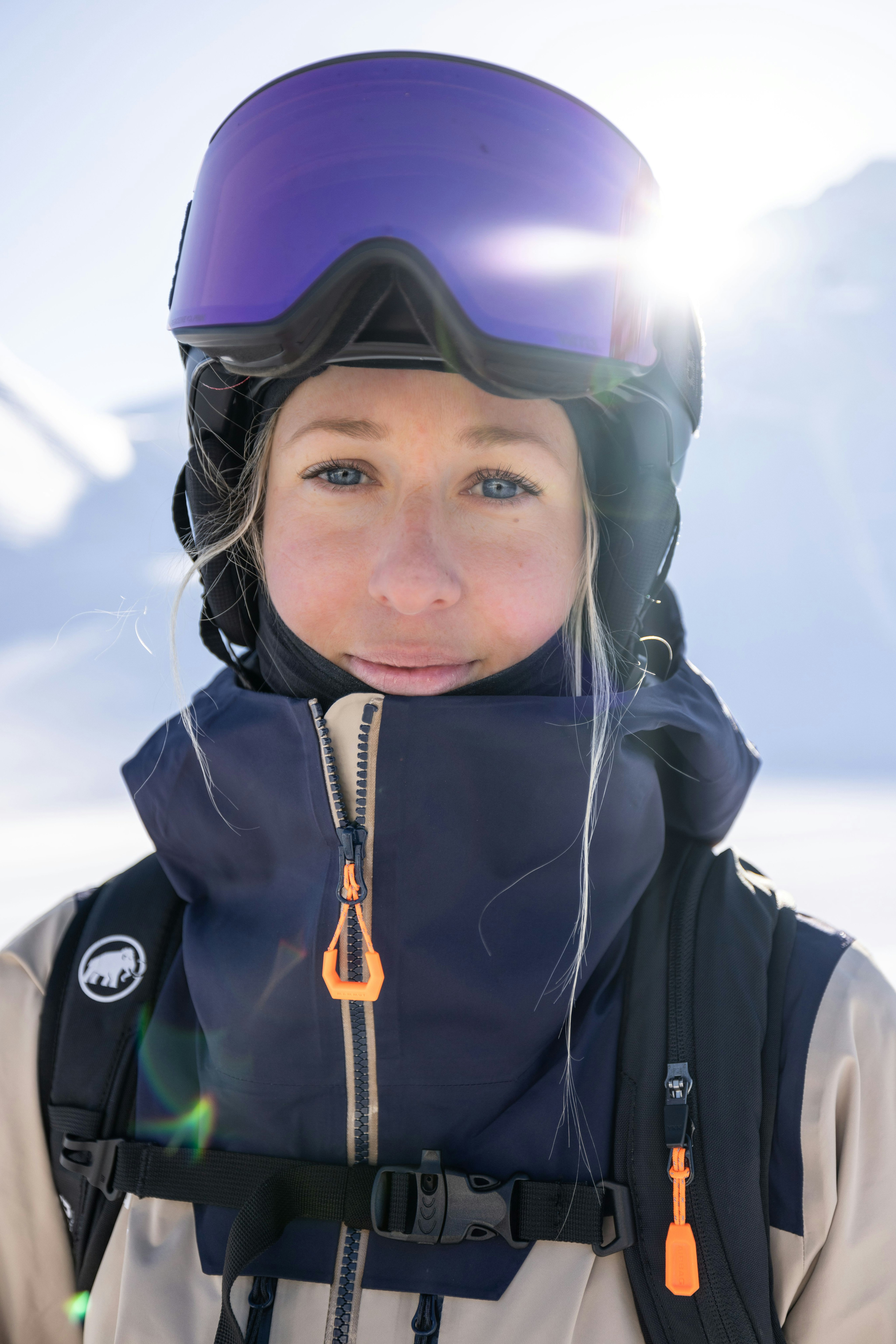 Linda in Mammut winter gear and goggles standing outdoors, snowy mountain range in the background with sunlight shining.