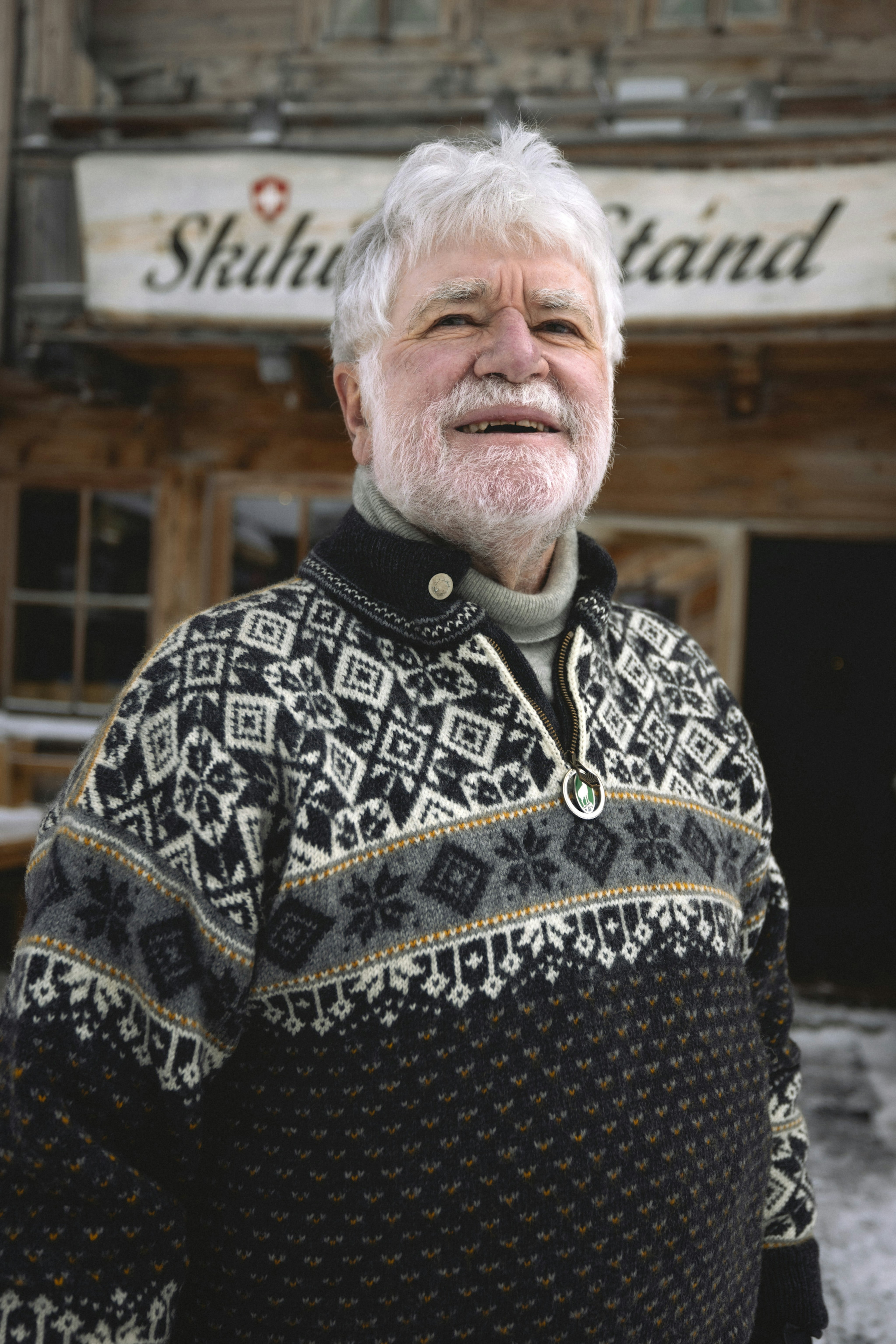 Elderly man in a Mammut patterned sweater smiling outdoors in front of a rustic wooden building.