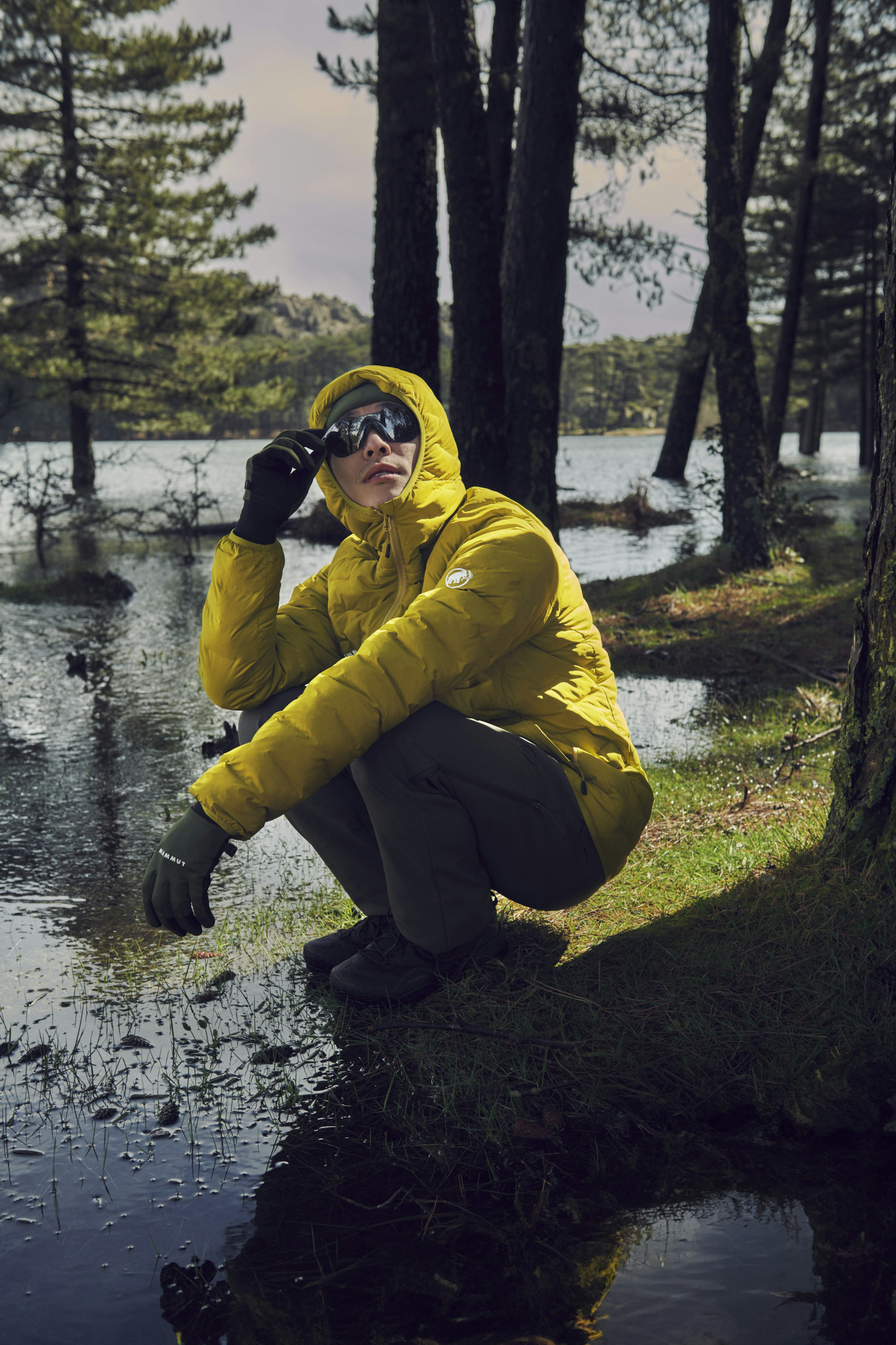 Person in a yellow Mammut jacket and sunglasses crouches near a forest pond, surrounded by trees, under a partly cloudy sky.