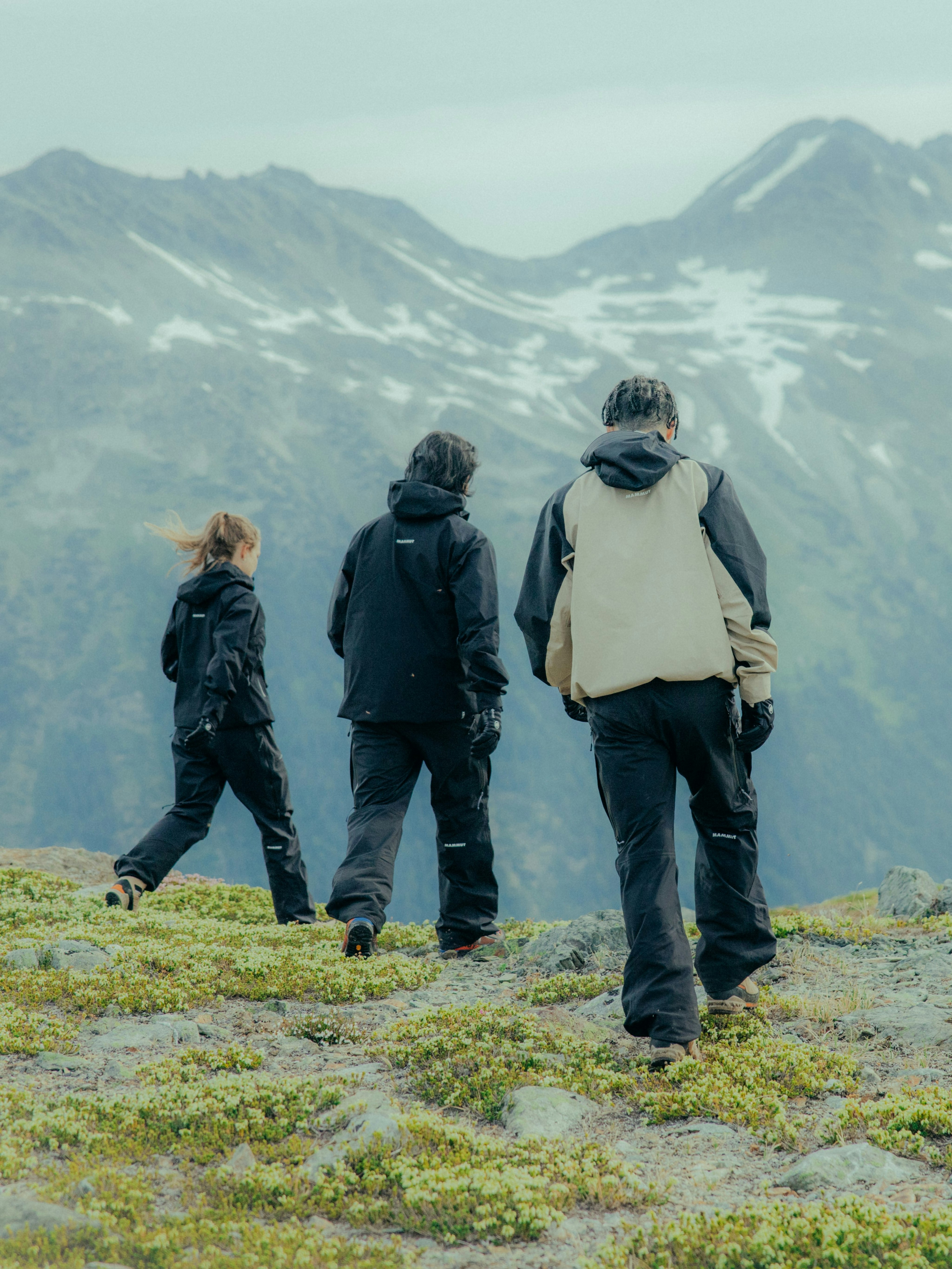 Three hikers in black Mammut outdoor gear trekking up a grassy hill with stunning snow-capped mountains in the background.