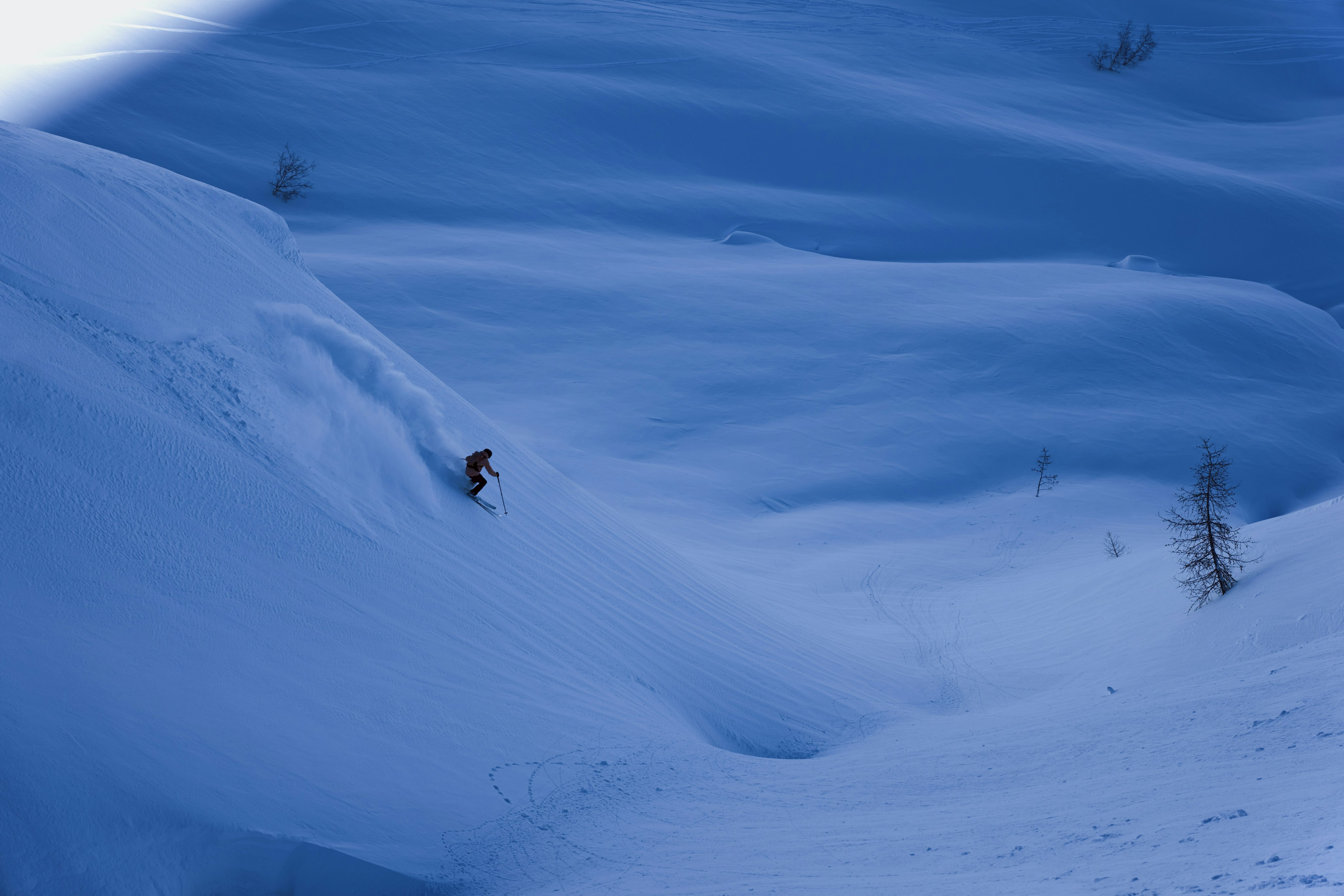 A skier wearing Mammut gear expertly descends a steep, snowy slope under a clear blue sky, carving through fresh powder and leaving an impressive trail behind.