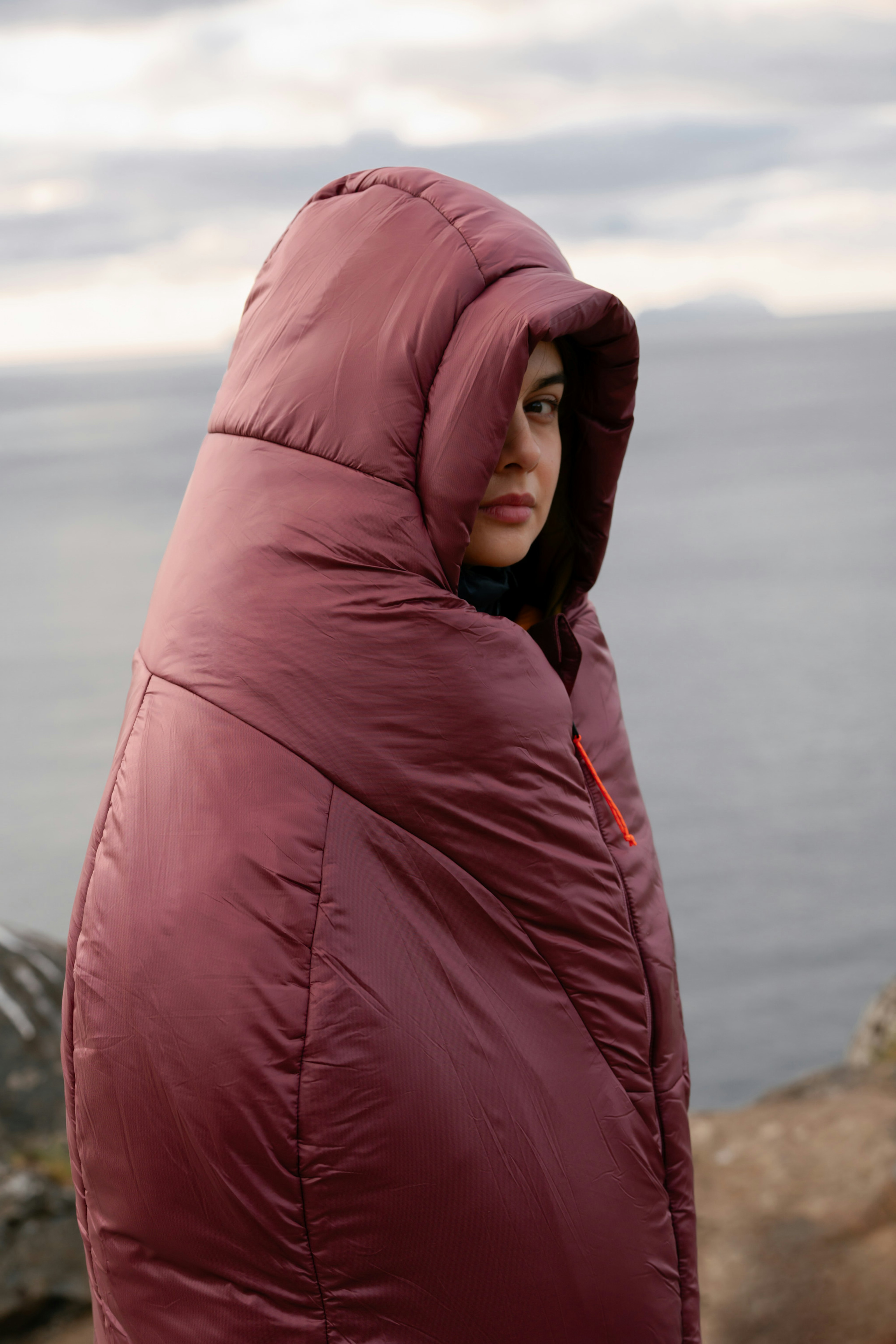 Person wrapped in a red Mammut sleeping bag, standing outdoors near a lake with a cloudy sky in the background.