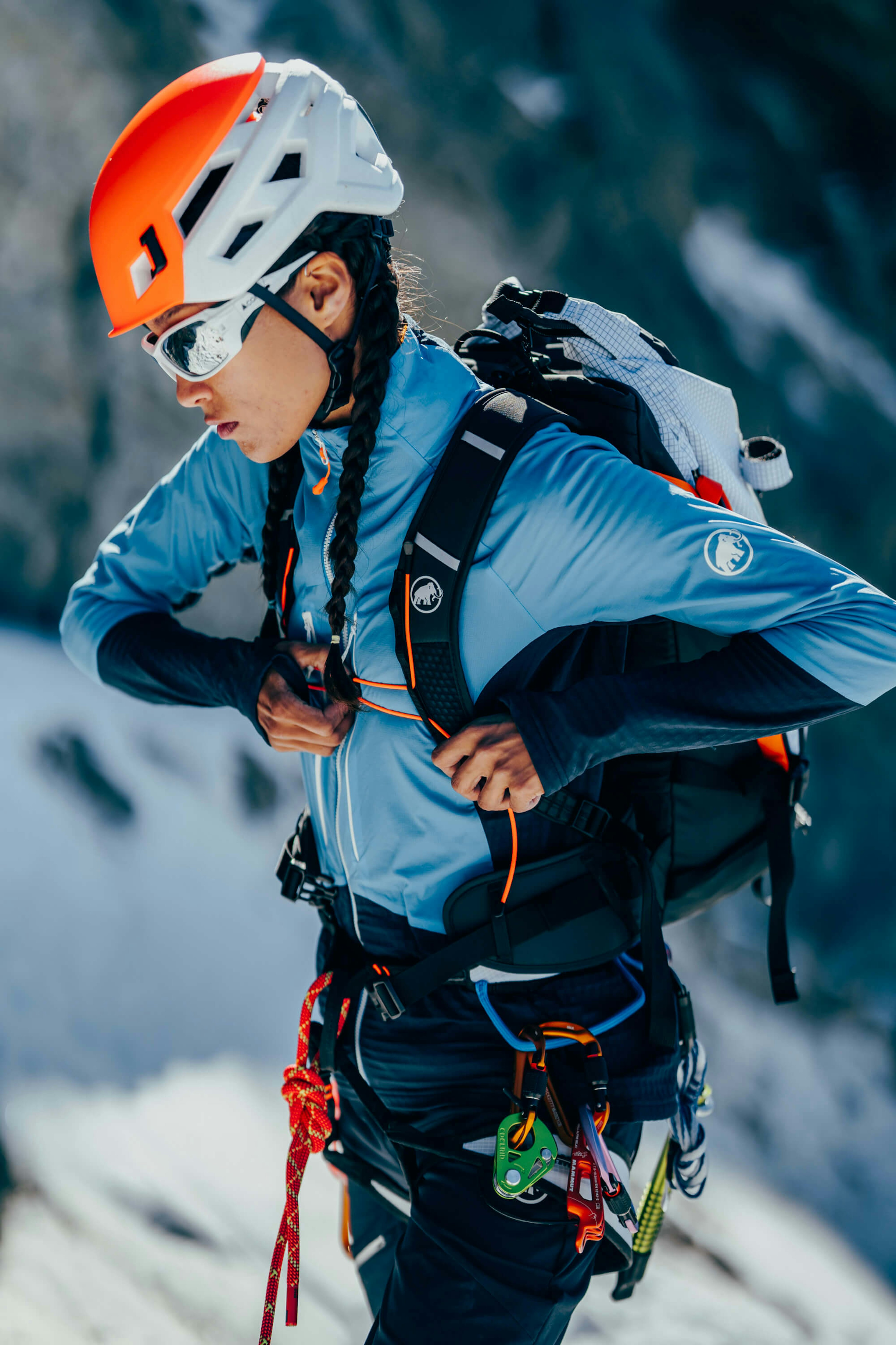 A Mammut climber wearing a helmet, sunglasses, and premium climbing gear adjusts harness straps on snowy mountain terrain.