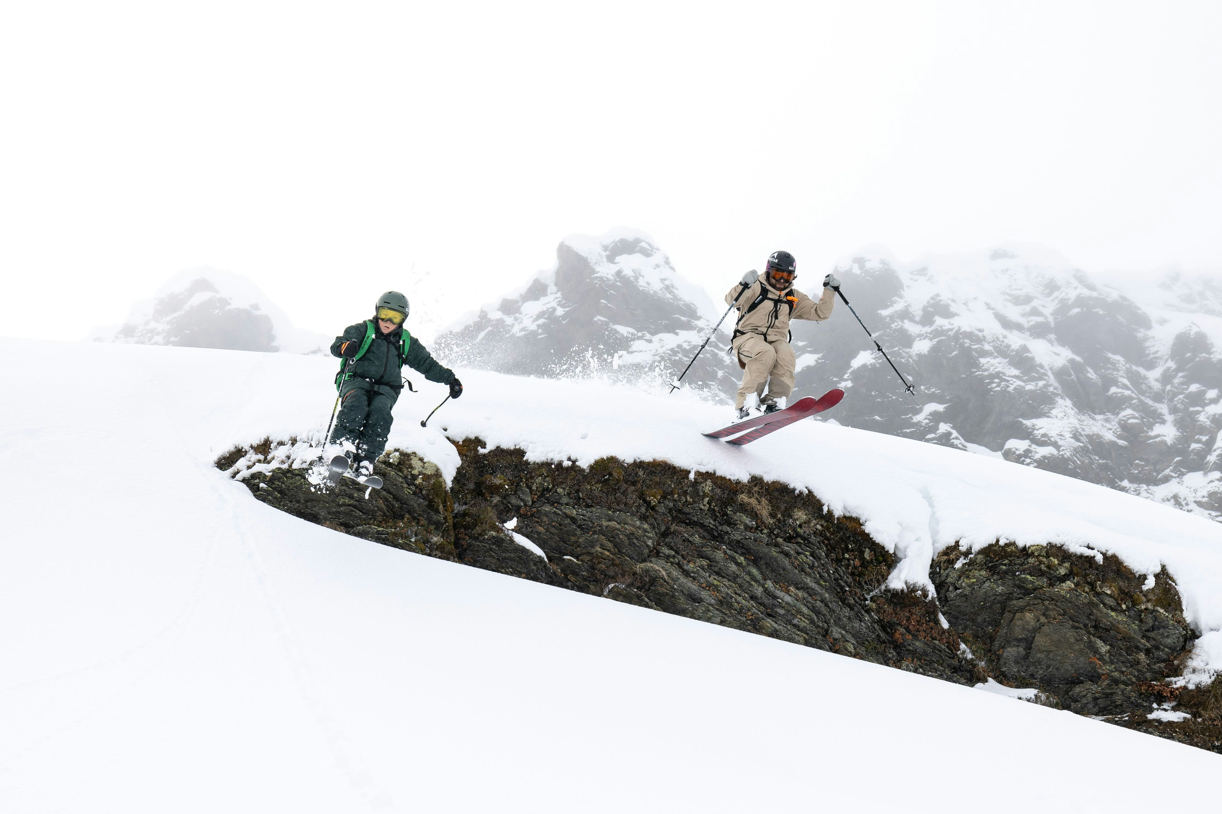 Whisky and his son, two skiers in vibrant Mammut gear navigate a snowy mountain with rocky patches, beneath a cloudy sky.