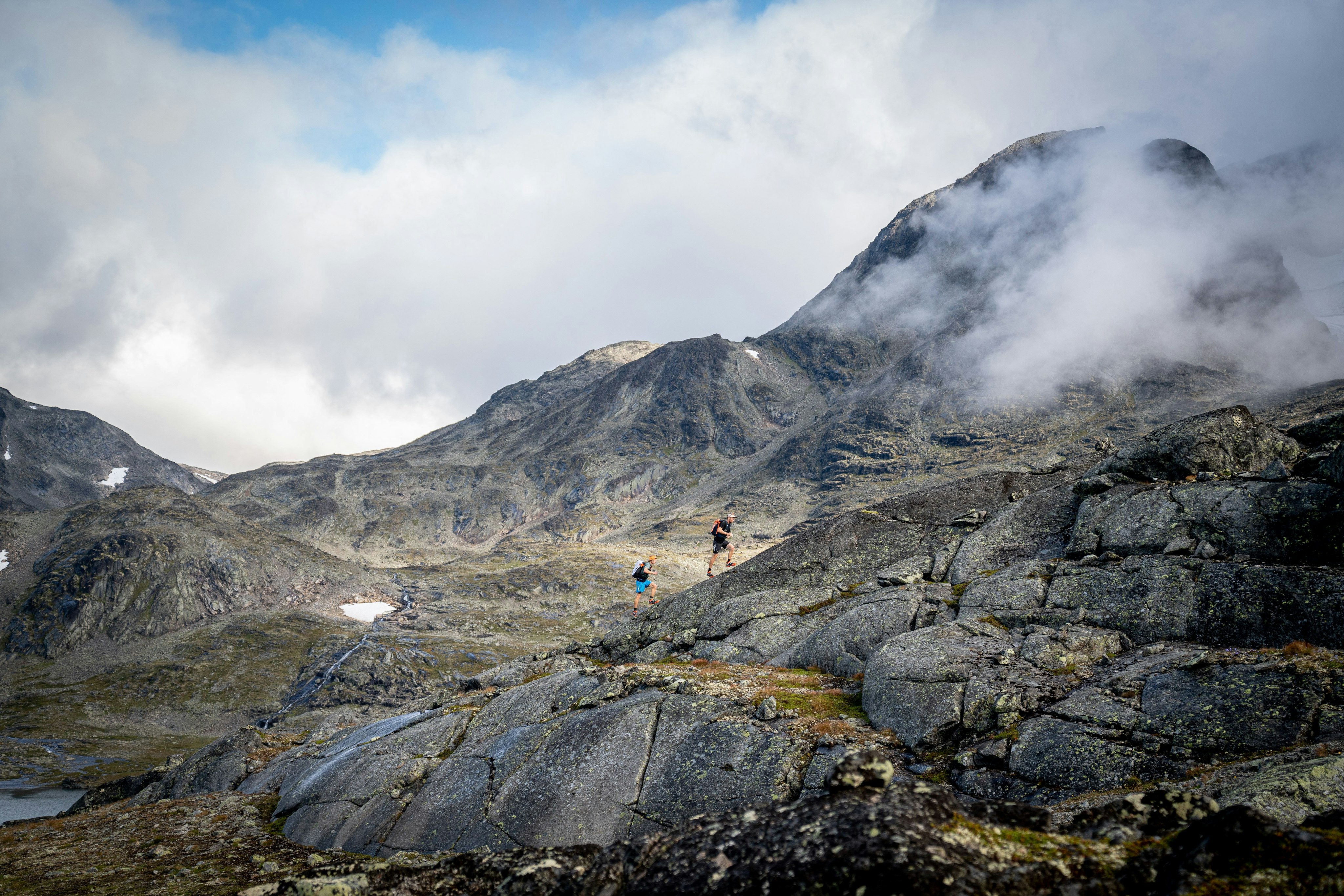 Two hikers equipped with Mammut gear traverse rugged, rocky terrain in a mist-covered mountainous landscape under a partly cloudy sky.