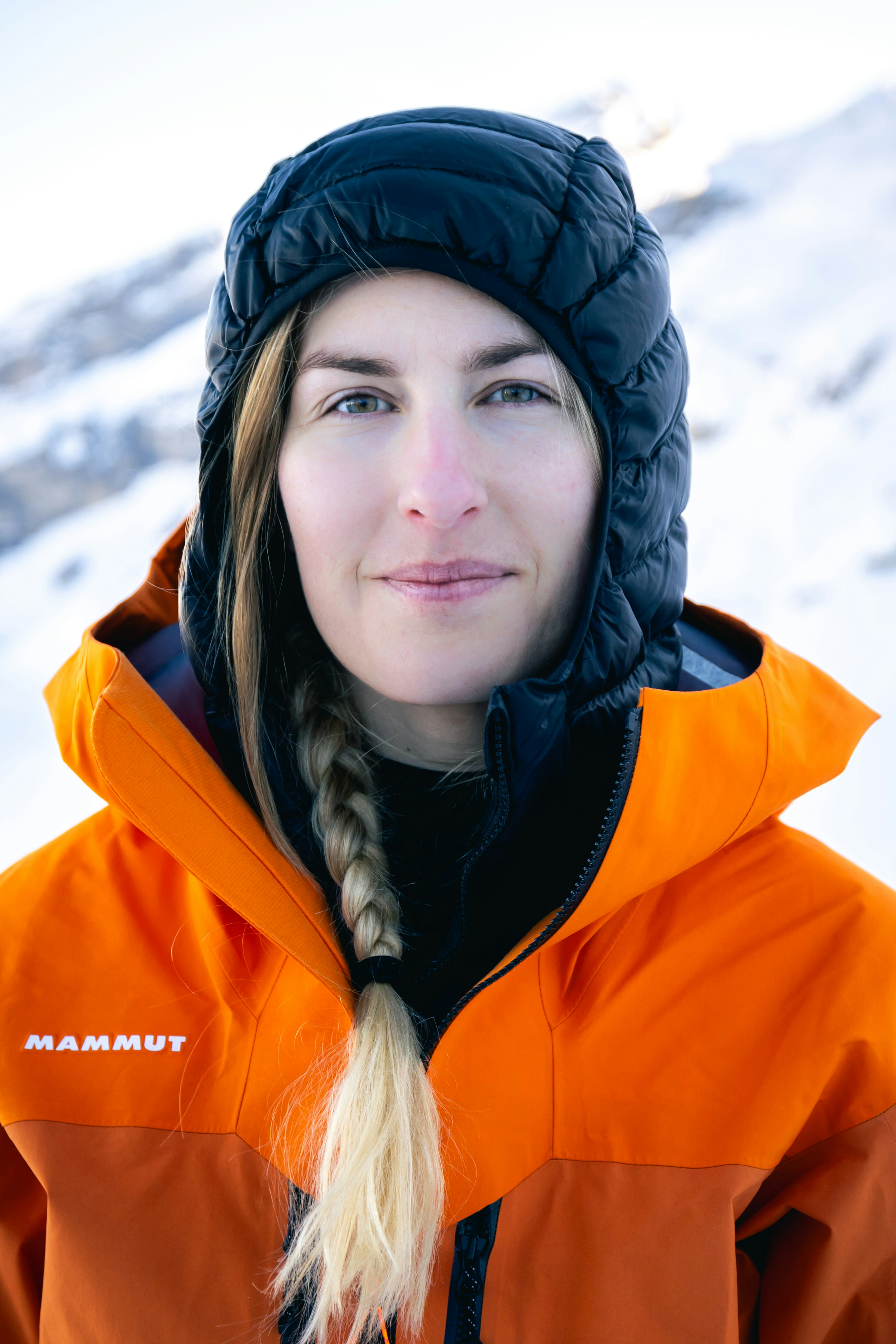 Person wearing a Mammut orange jacket with a black hood, standing outdoors in a snowy mountain landscape.