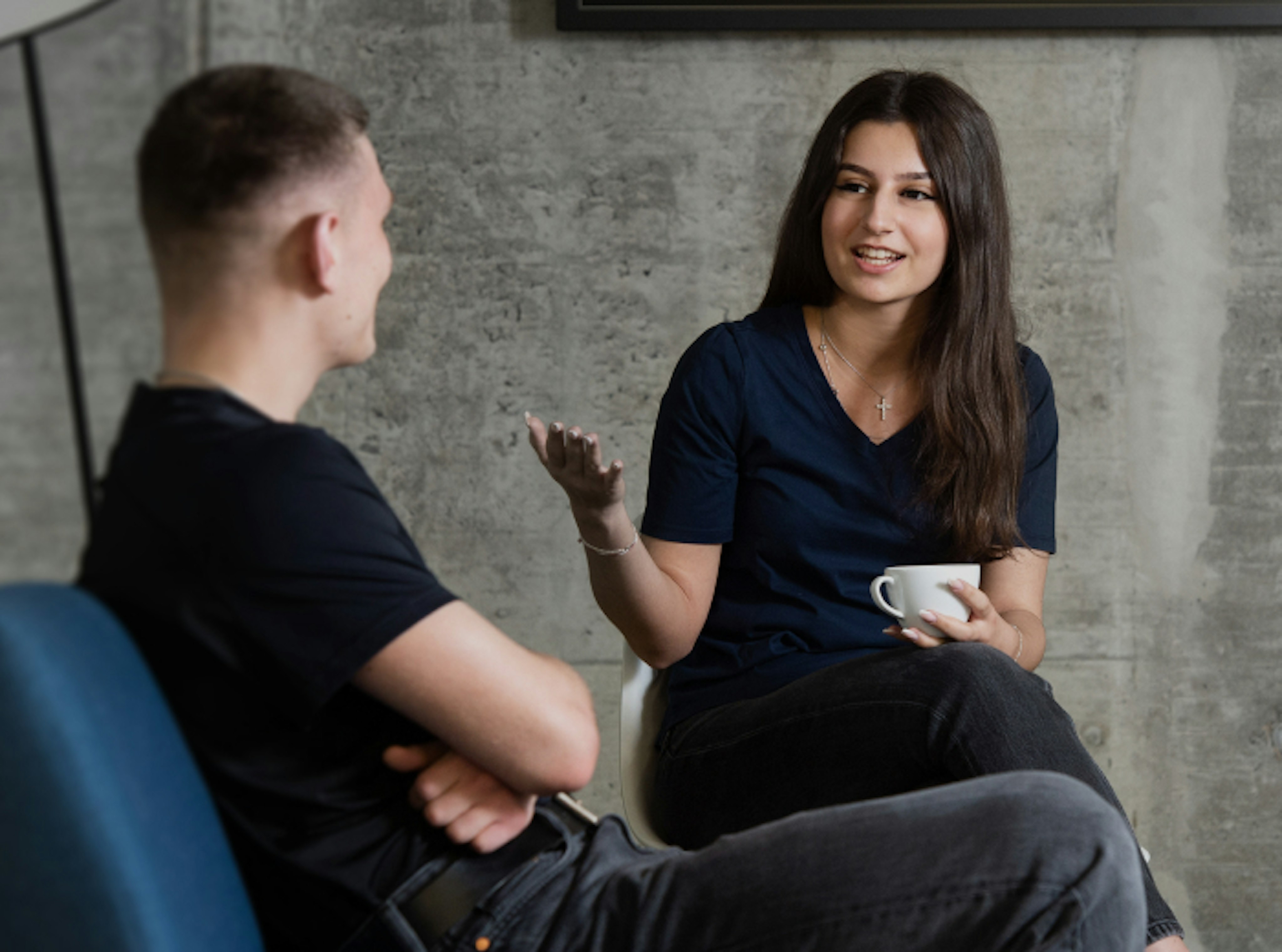 Two people engaged in a conversation, one holding a coffee cup, sitting on Mammut-branded chairs against a gray concrete wall.