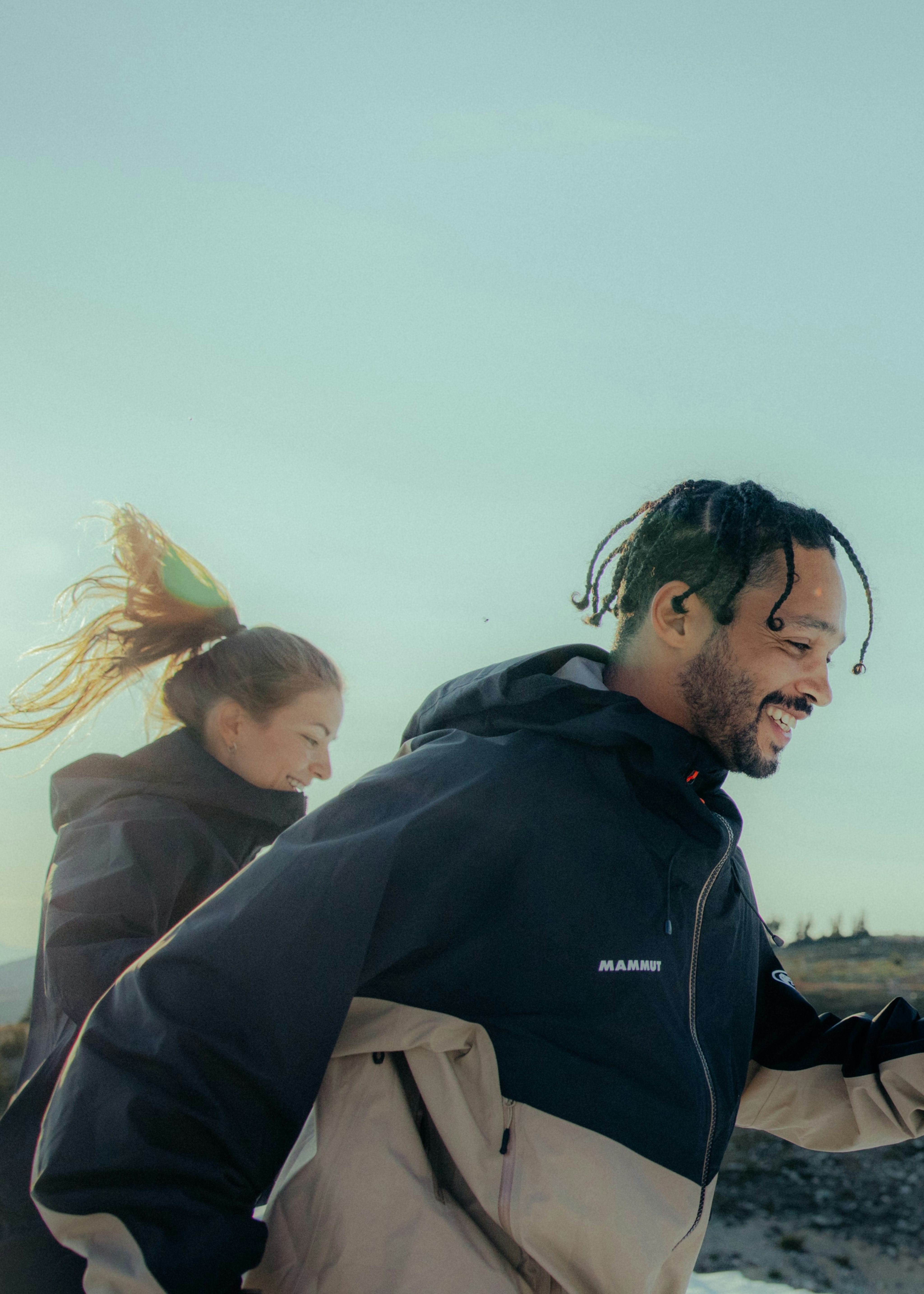 Two adventurous individuals in Mammut outdoor jackets smiling and running beneath a light sky background.