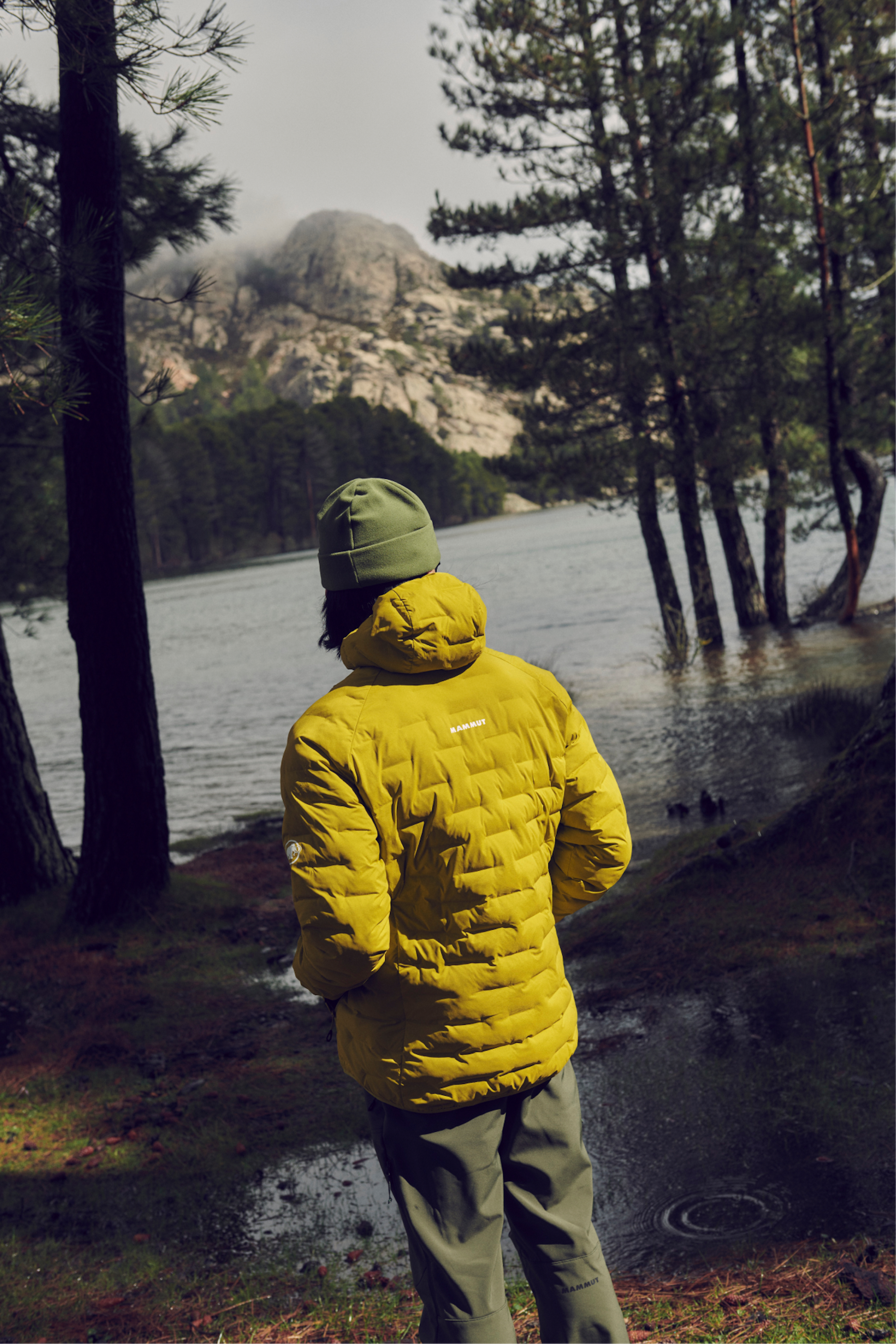 Person wearing a yellow Mammut jacket and green beanie stands by a serene lake, surrounded by lush trees and majestic mountains in the background.