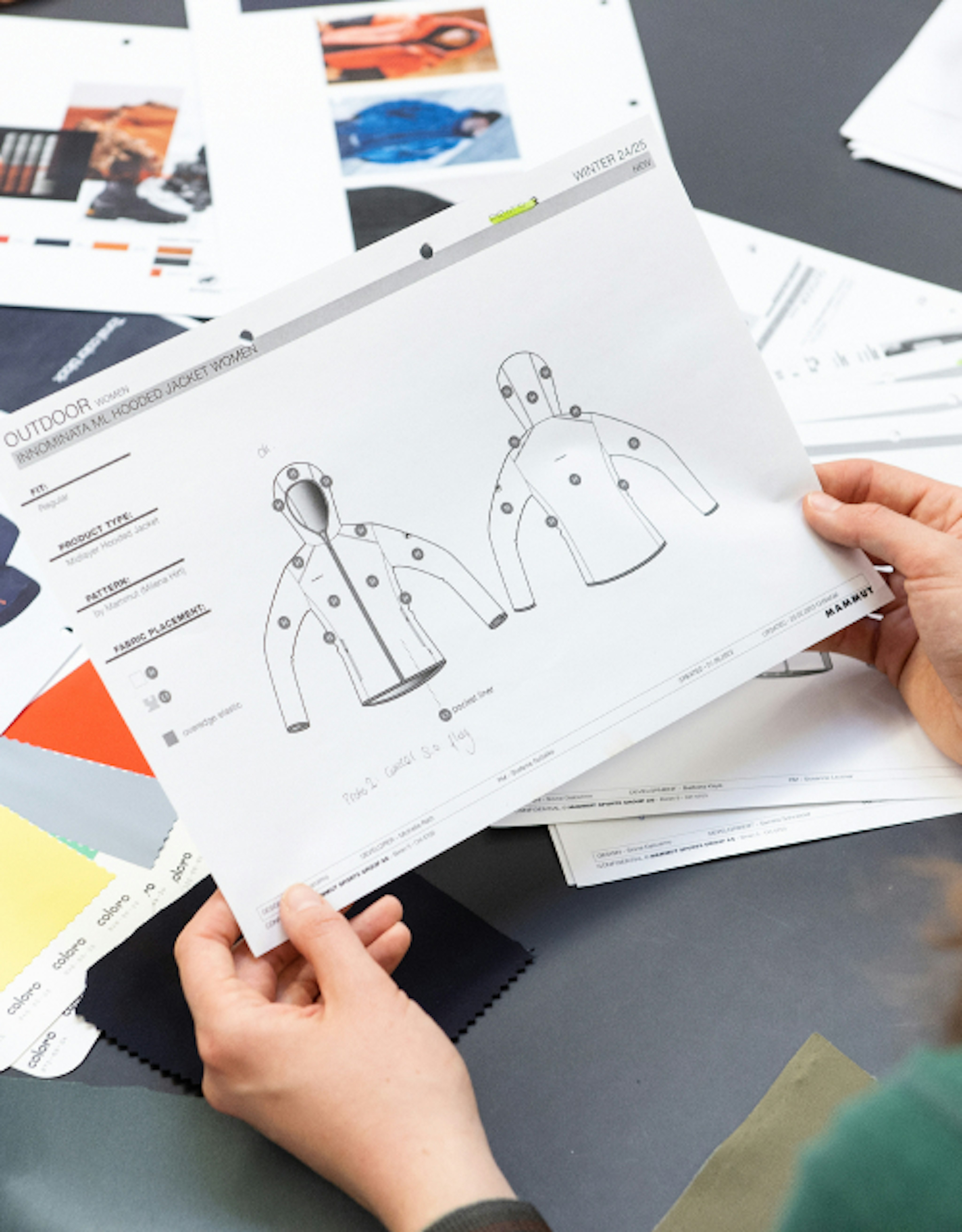 Close-up of hands holding a Mammut jacket design sheet with fabric swatches and design documents on a table.