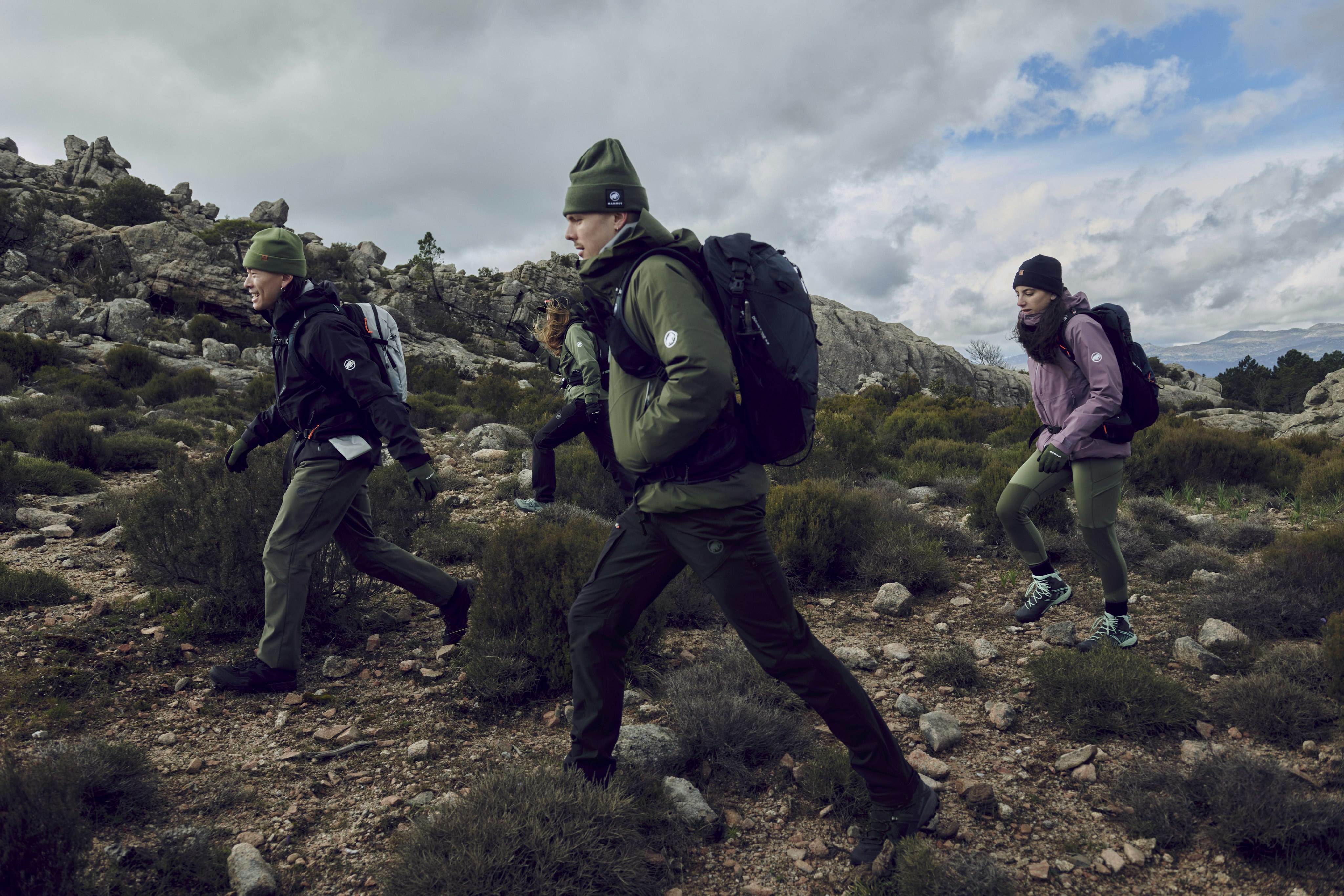 A group of hikers in Mammut winter clothing, carrying Mammut backpacks, navigate a rocky trail surrounded by majestic mountains and shrubs.