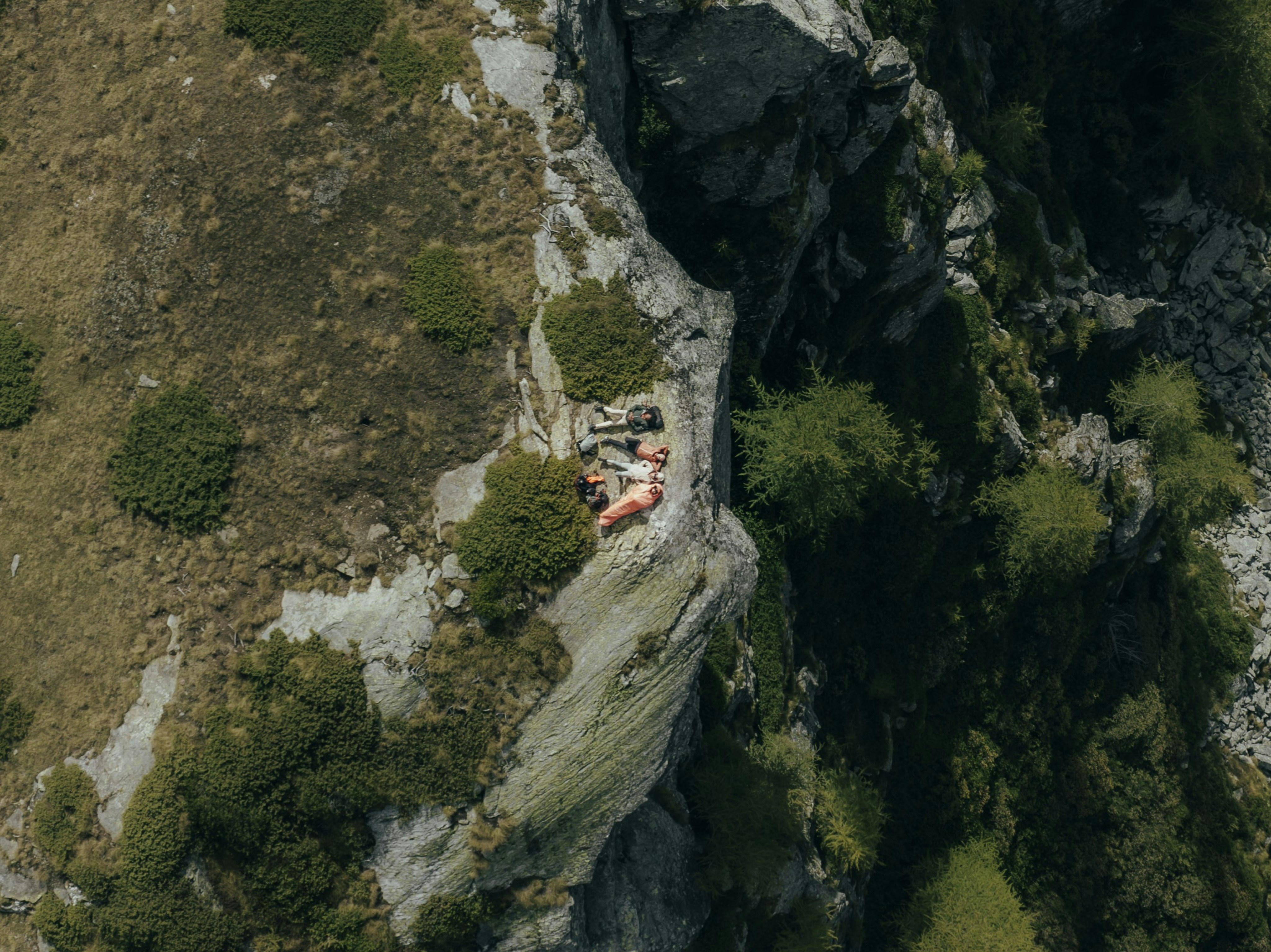 Person wearing Mammut gear resting on a rocky cliff ledge above a deep crevice, surrounded by sparse vegetation and rugged mountain terrain.