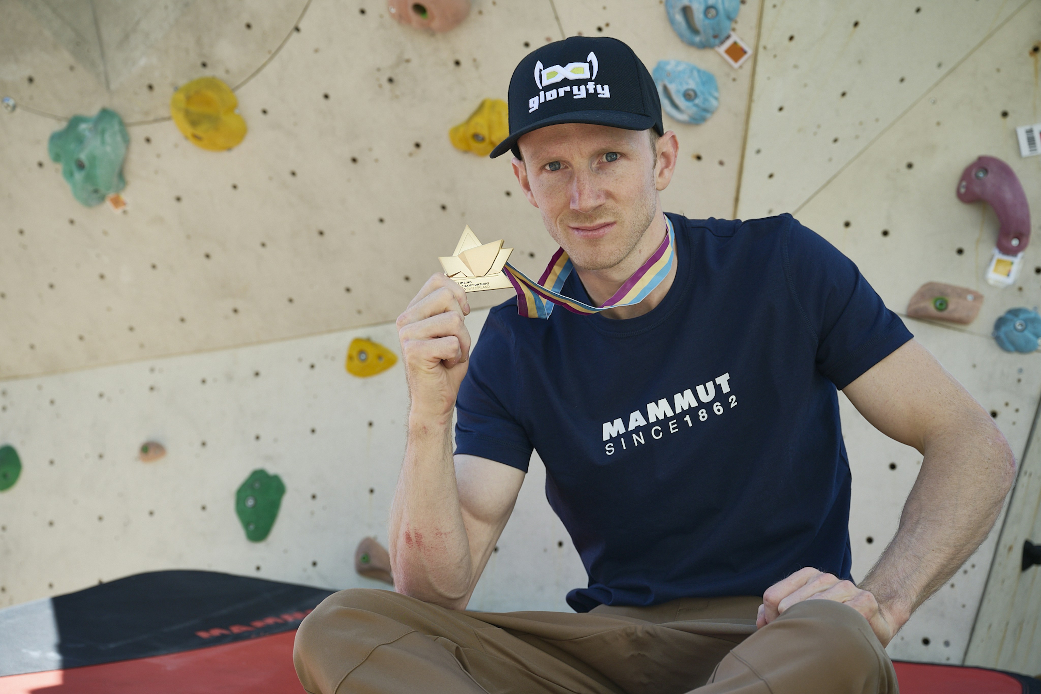 Jakob Schubert, a climber wearing a blue Mammut t-shirt and cap proudly displays a gold medal in front of an indoor climbing wall.