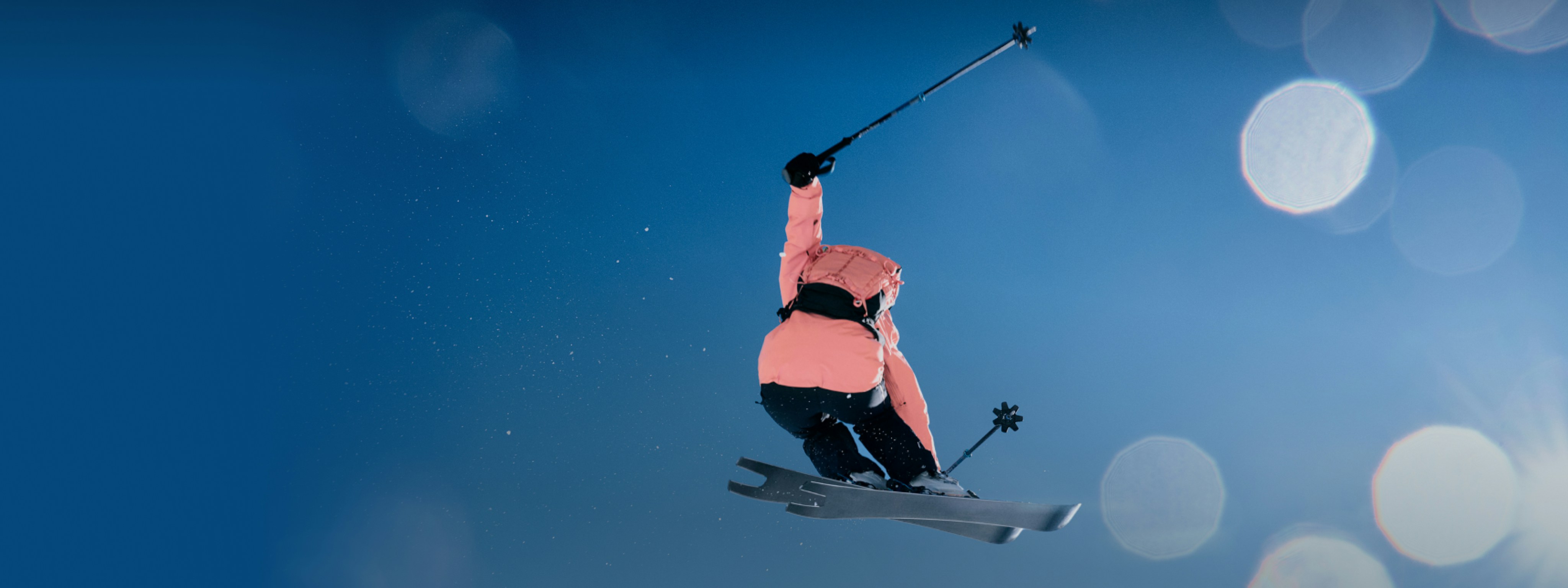 Skier in a Mammut pink Stoney jacket performing a mid-air stunt against a vibrant blue sky.