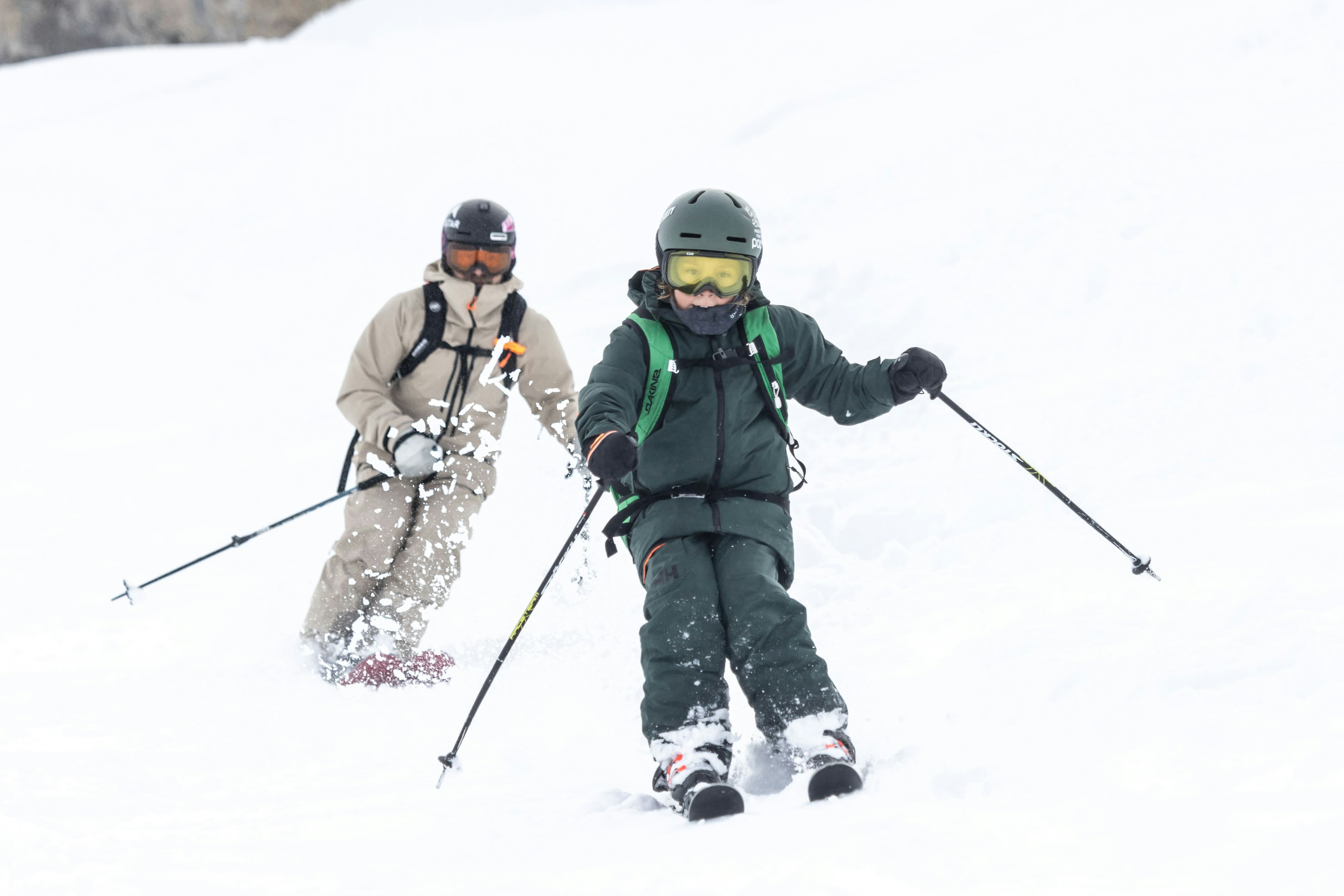 Whisky and his son in Mammut winter gear expertly carving down a snow-covered slope, with powdery snow dramatically flying up around them.