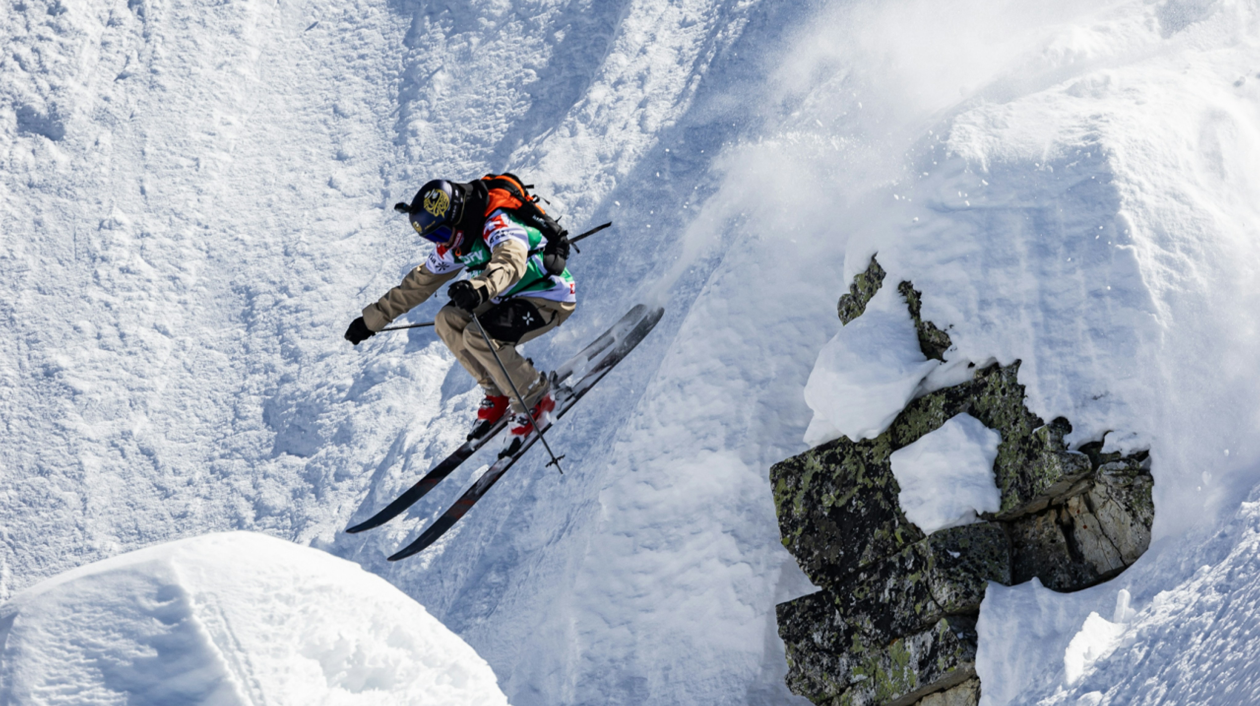 A Mammut skier in mid-air performing an impressive jump off a snowy cliff with stunning snow-covered mountains in the background.