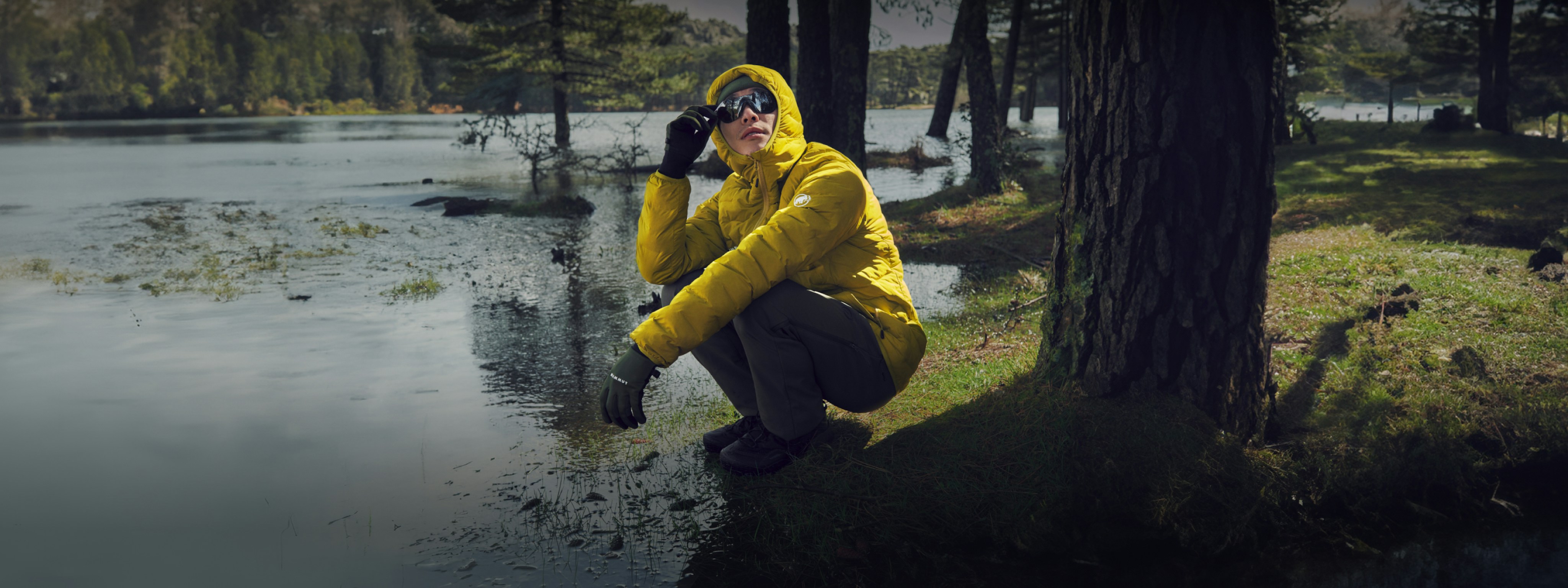 Person in a yellow Mammut jacket and sunglasses crouching beside a forest lake, with trees and their reflections in the water in the background.