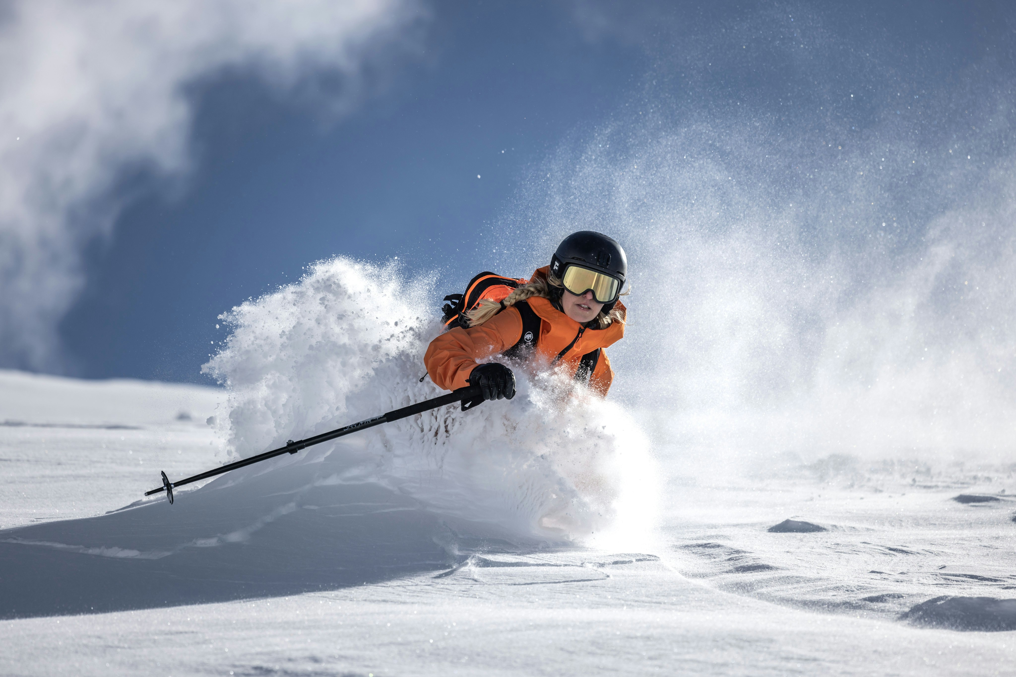 Sanne Mona wearing a Mammut orange jacket and black helmet effortlessly glides through pristine snowy terrain, leaving a trail of sparkling powder behind.